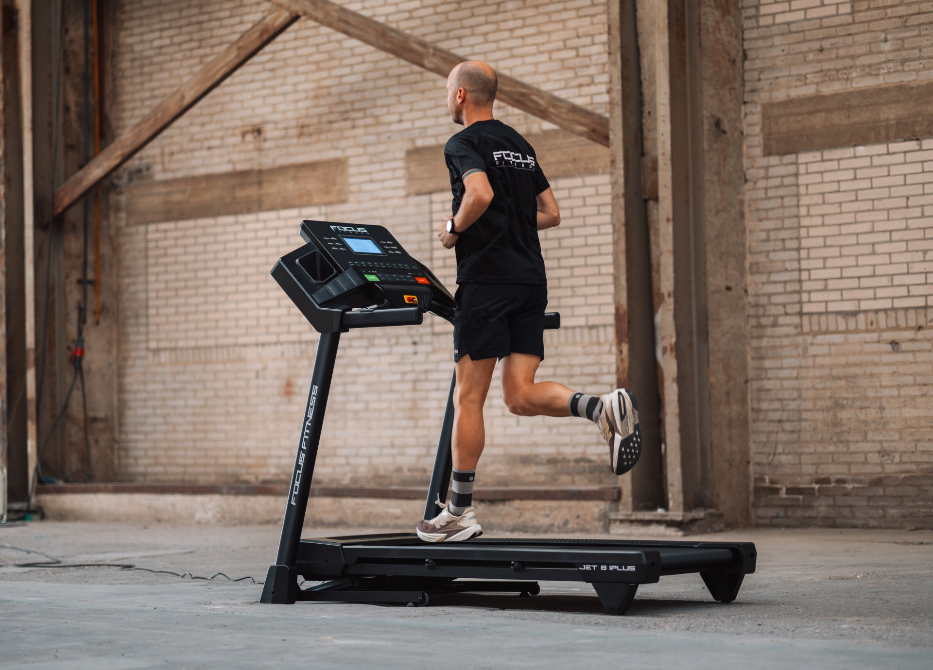A man runs on a treadmill in a brick-walled room. The treadmill is black, and the man wears black shorts and a shirt.