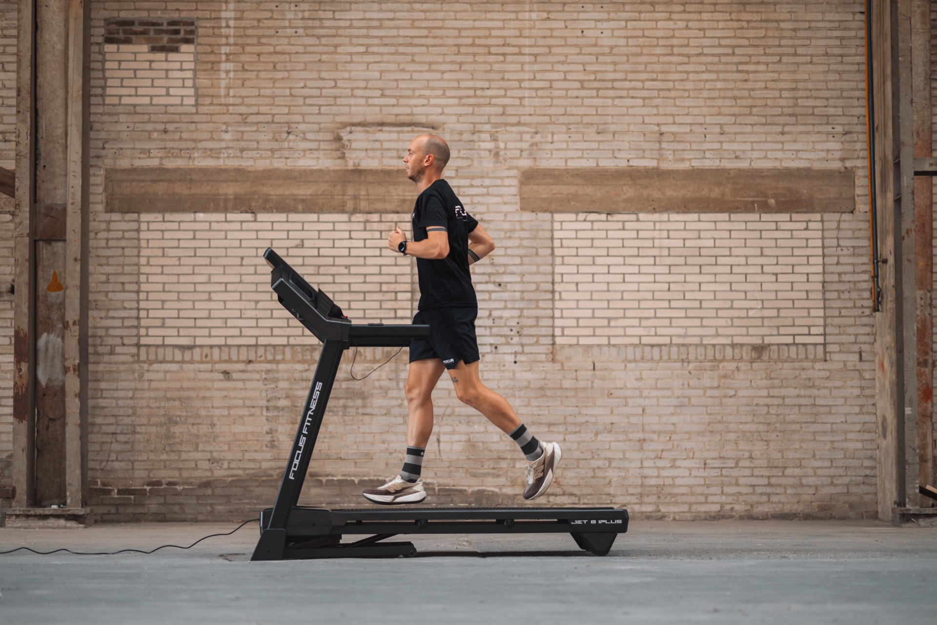 A man runs on a treadmill in an industrial space. He wears black shorts and a shirt. The background is a brick wall.