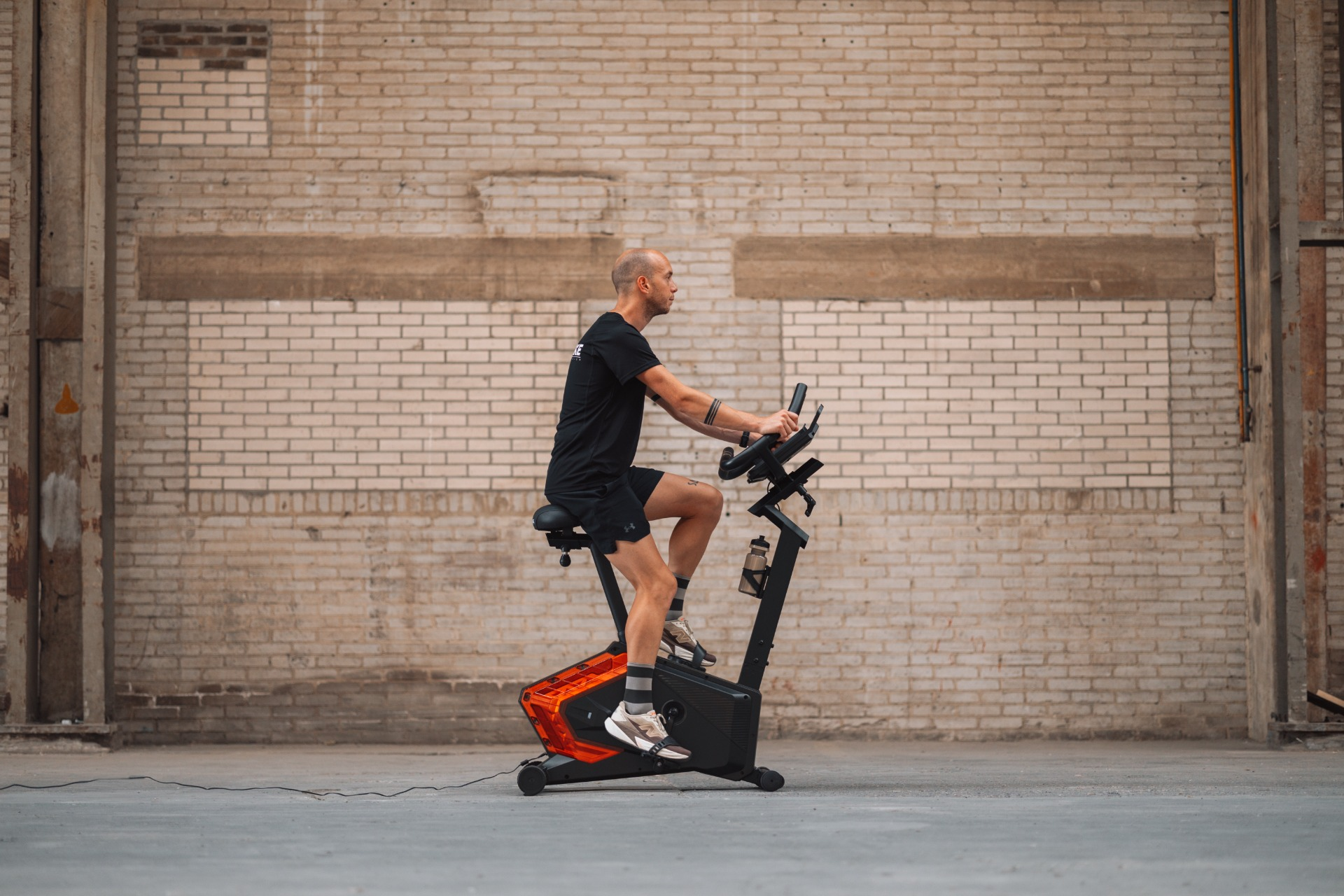 Een man in een zwarte korte broek en shirt fietst op een hometrainer. Achter hem is een bakstenen muur.