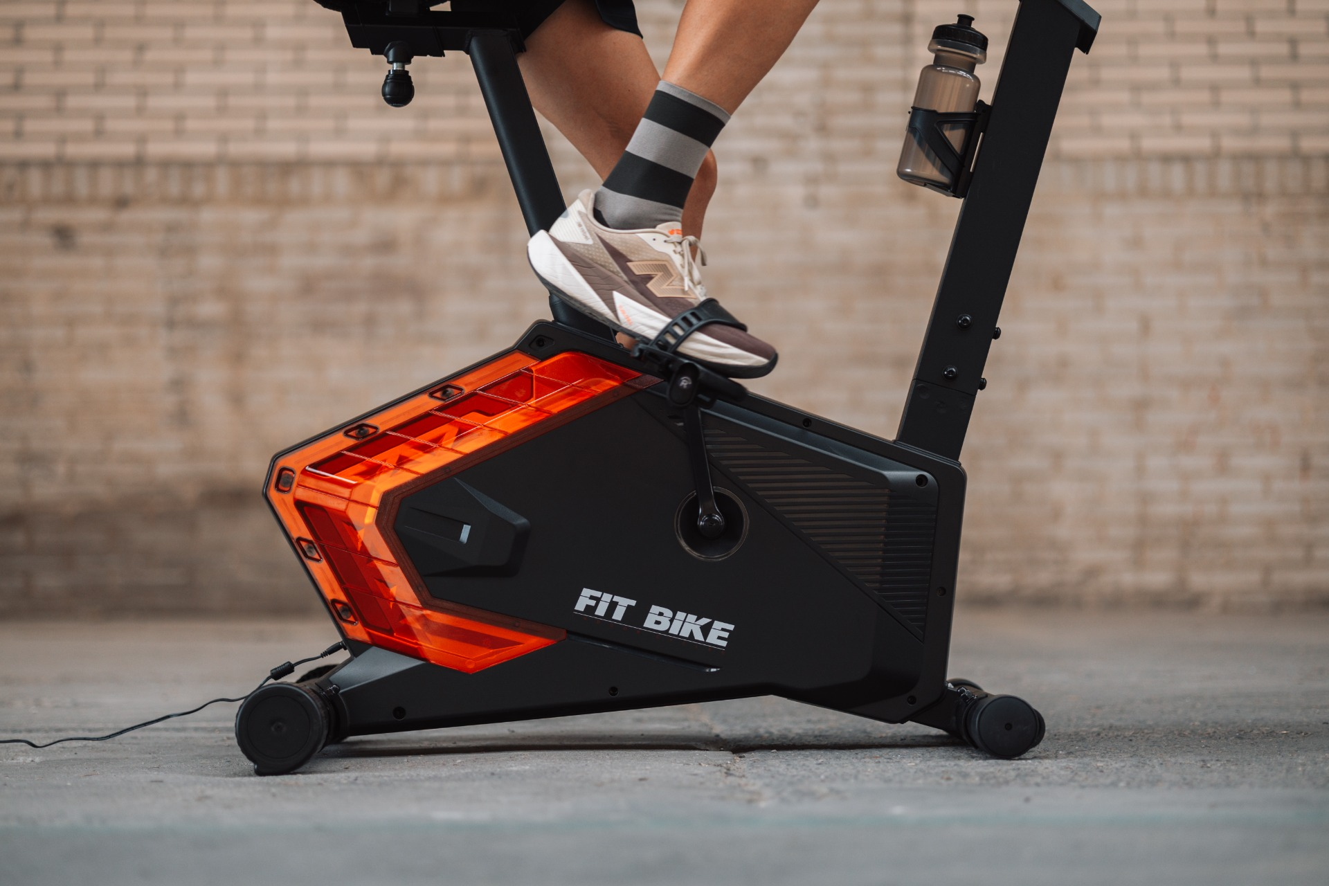 Been van een persoon op een Fit Bike. De fiets is zwart met oranje details. Een waterfles staat op het frame.