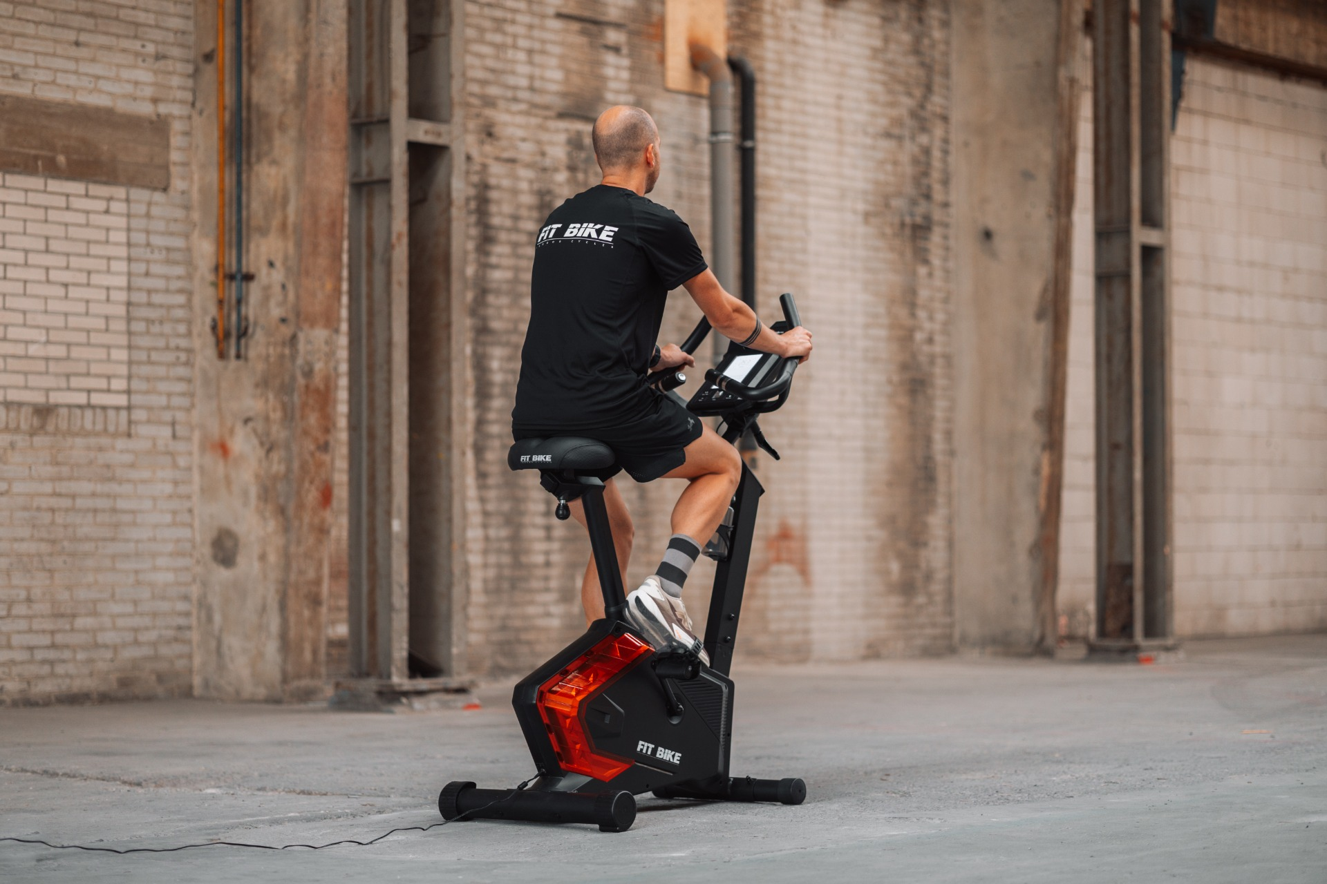 Een man op een hometrainer, achteraanzicht. De fiets is zwart en rood. De achtergrond is een bakstenen muur.