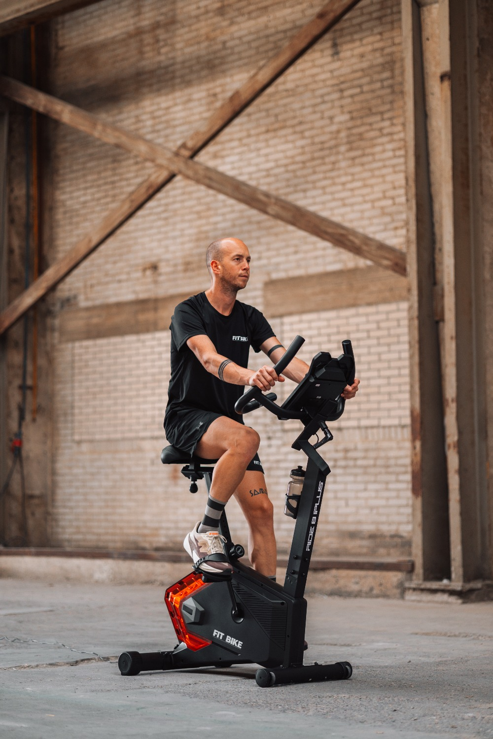 Man op een hometrainer in een magazijn. Hij draagt zwart, met een tatoeage. De fiets heeft rode lichten. Bakstenen muur op de achtergrond.