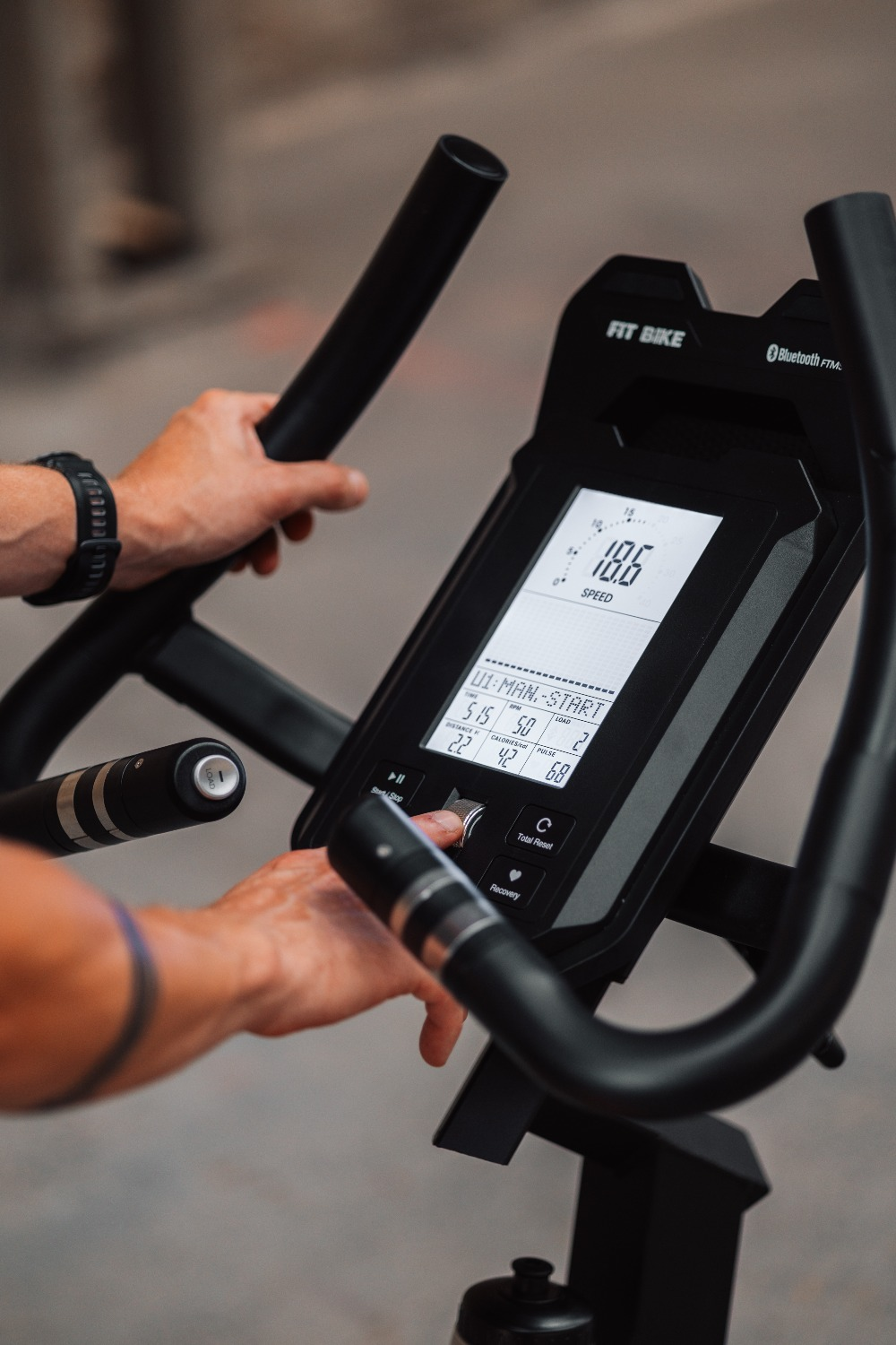 Een persoon gebruikt een Fit Bike. Het display toont snelheid en andere statistieken. Handen zijn op de handvatten en passen de bediening aan.