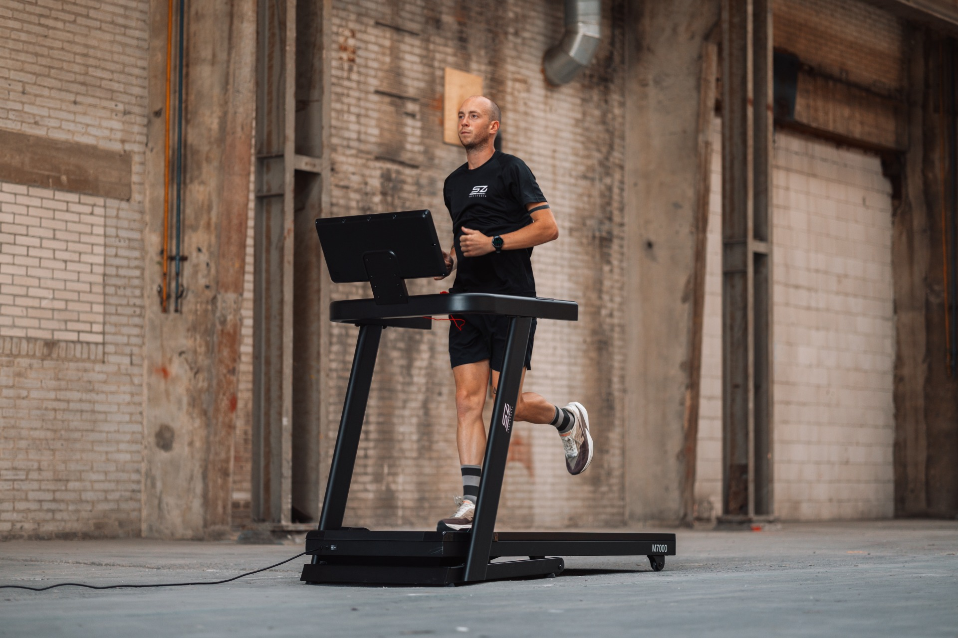 A man runs on a treadmill in a warehouse. He wears a black shirt and shorts. The treadmill is black.