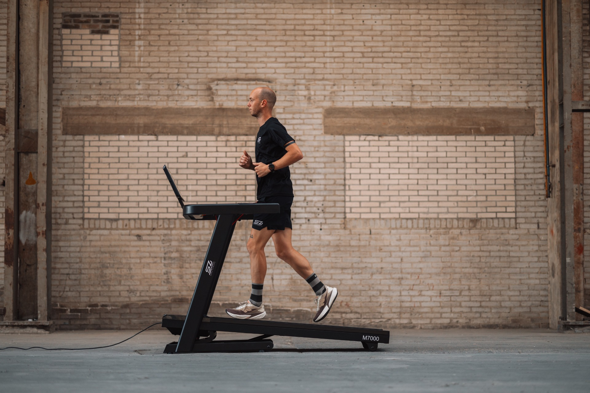 A man runs on a treadmill in a warehouse. He wears black athletic wear. The background is a brick wall.