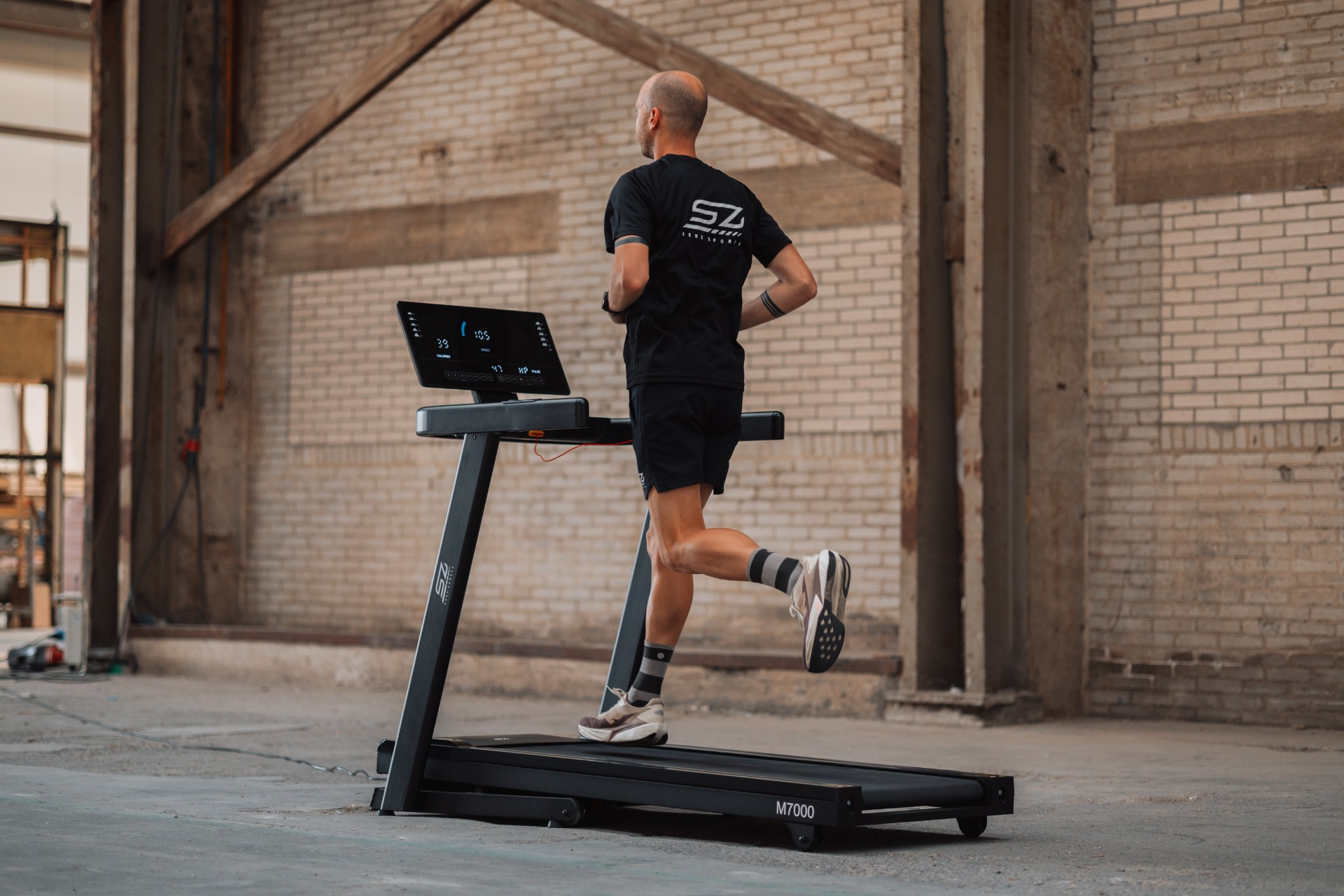 A man runs on a treadmill in a warehouse. He wears black clothing and shoes. The treadmill displays running data.
