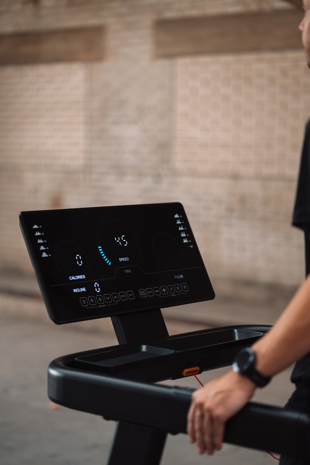 A treadmill display shows 4.5 speed. A person is touching the handrail. The background is a brick wall.