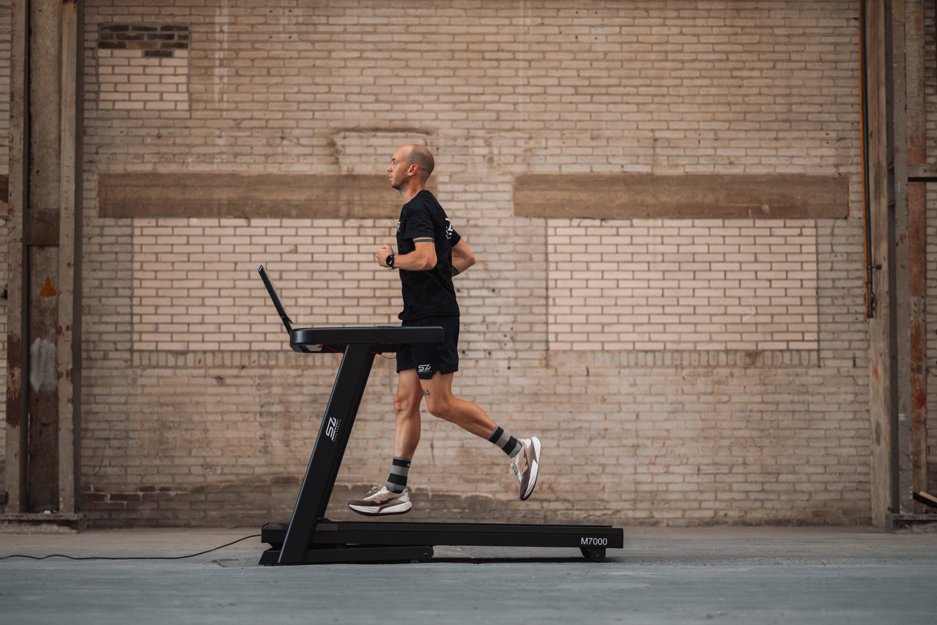 A man runs on a treadmill in a brick-walled space. He wears black shorts, shirt, and running shoes.