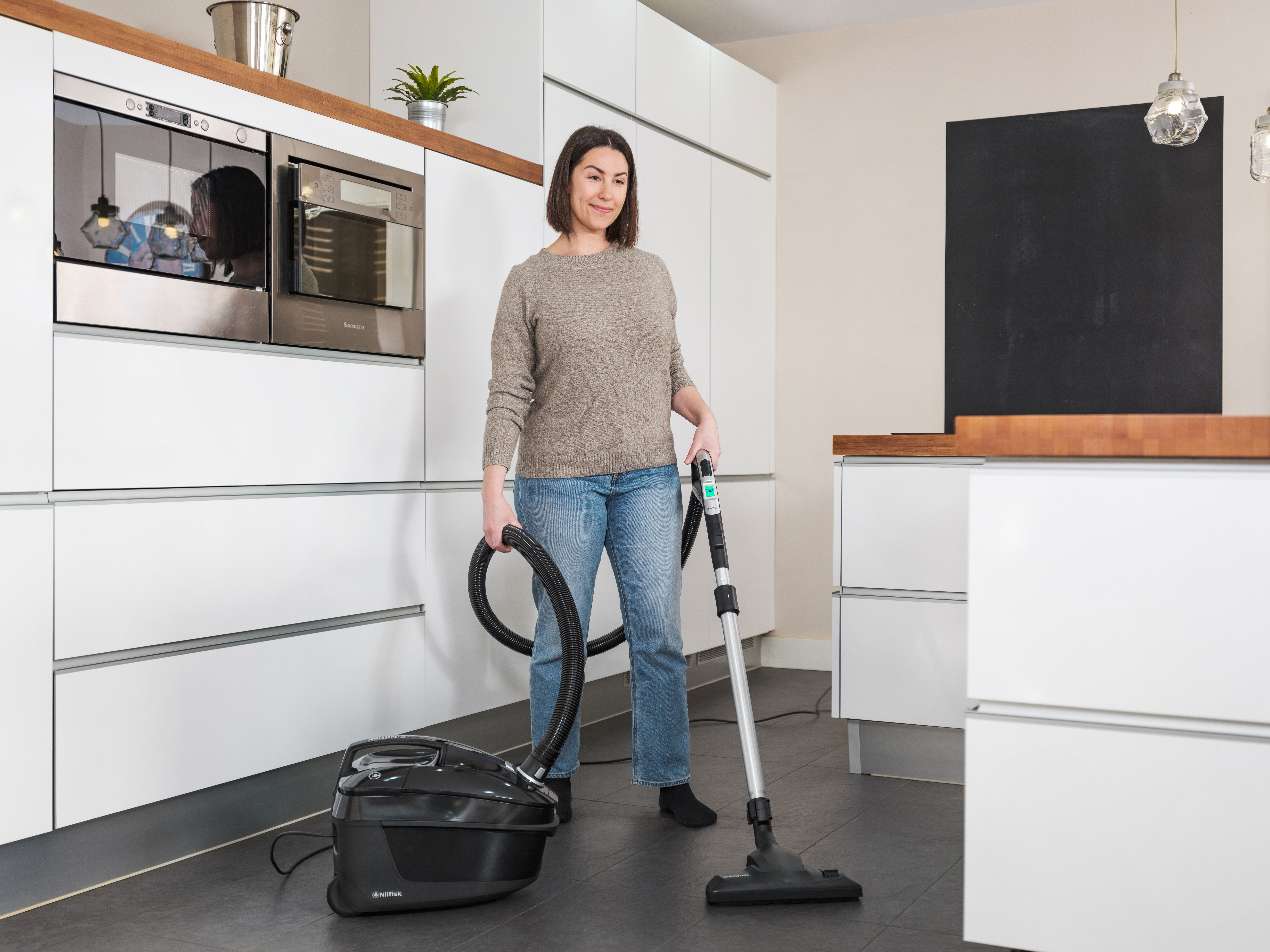 Kobieta w kuchni odkurza podłogę czarnym odkurzaczem. Kuchnia ma białe szafki i sprzęty.