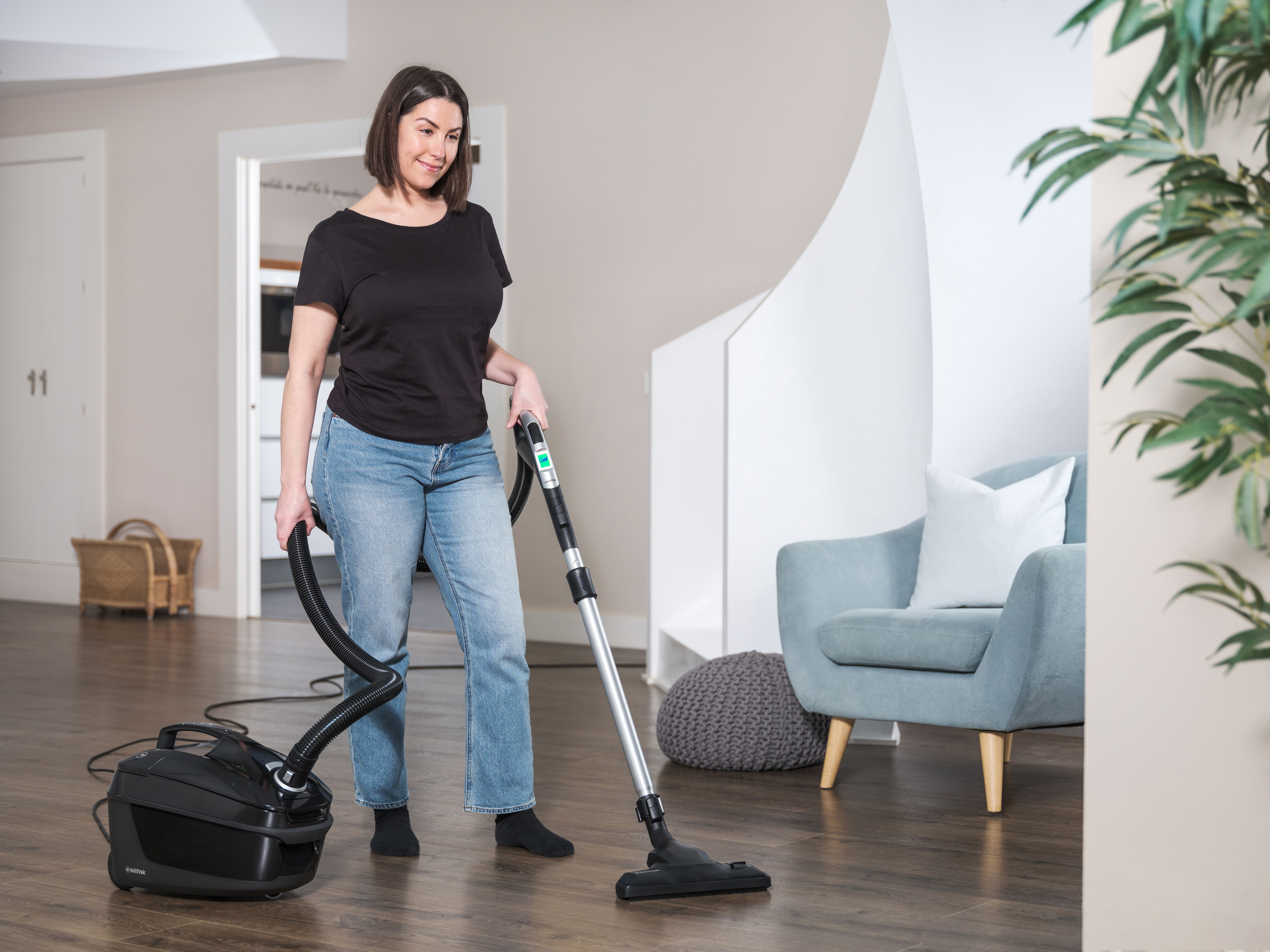 Kobieta odkurza drewnianą podłogę w domu. Ma na sobie czarną koszulę i dżinsy. Szary fotel i roślina w tle.