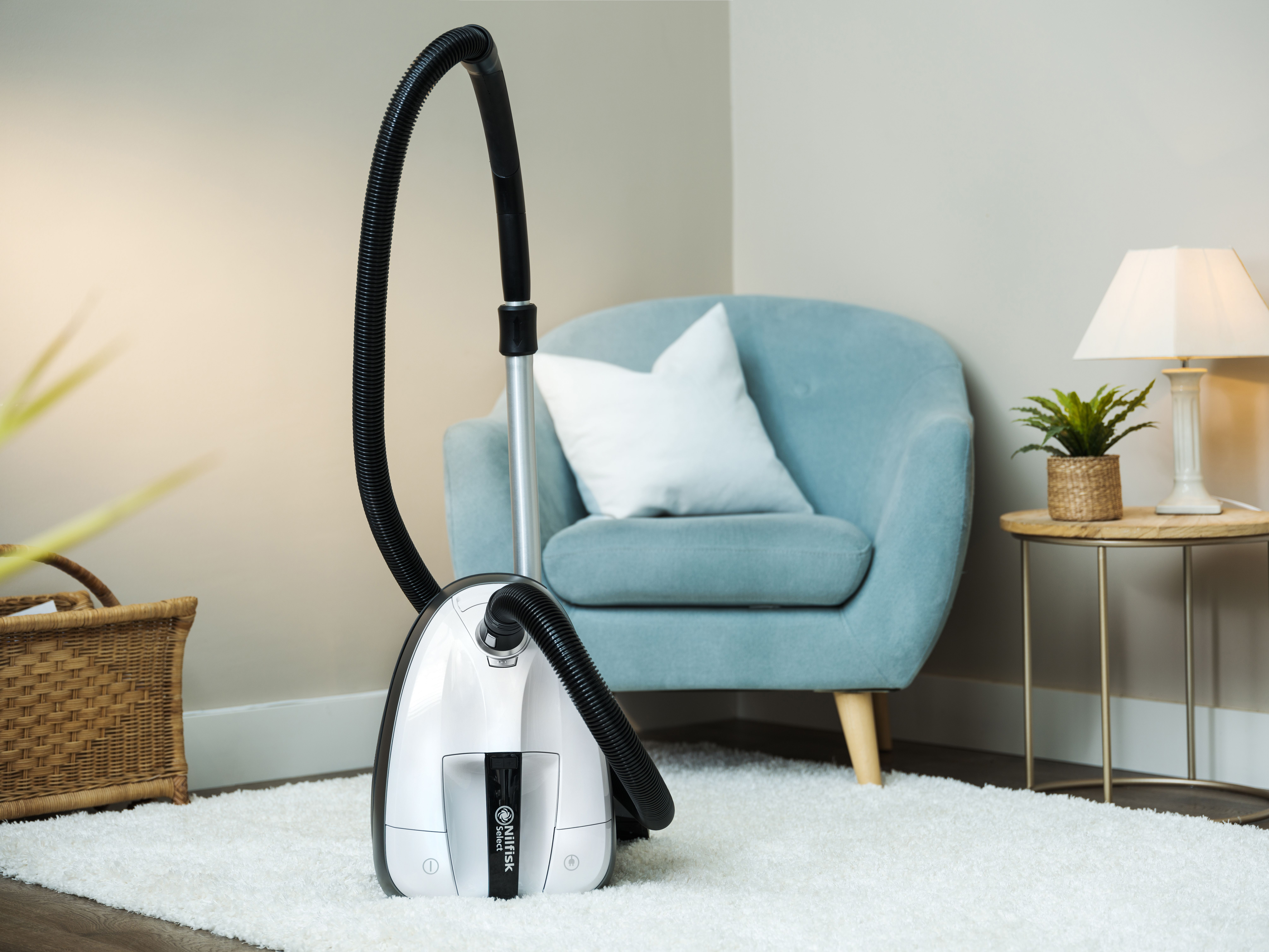 Un aspirateur blanc est sur un tapis blanc. Un fauteuil bleu, une table d'appoint avec une lampe et un panier sont en arrière-plan.