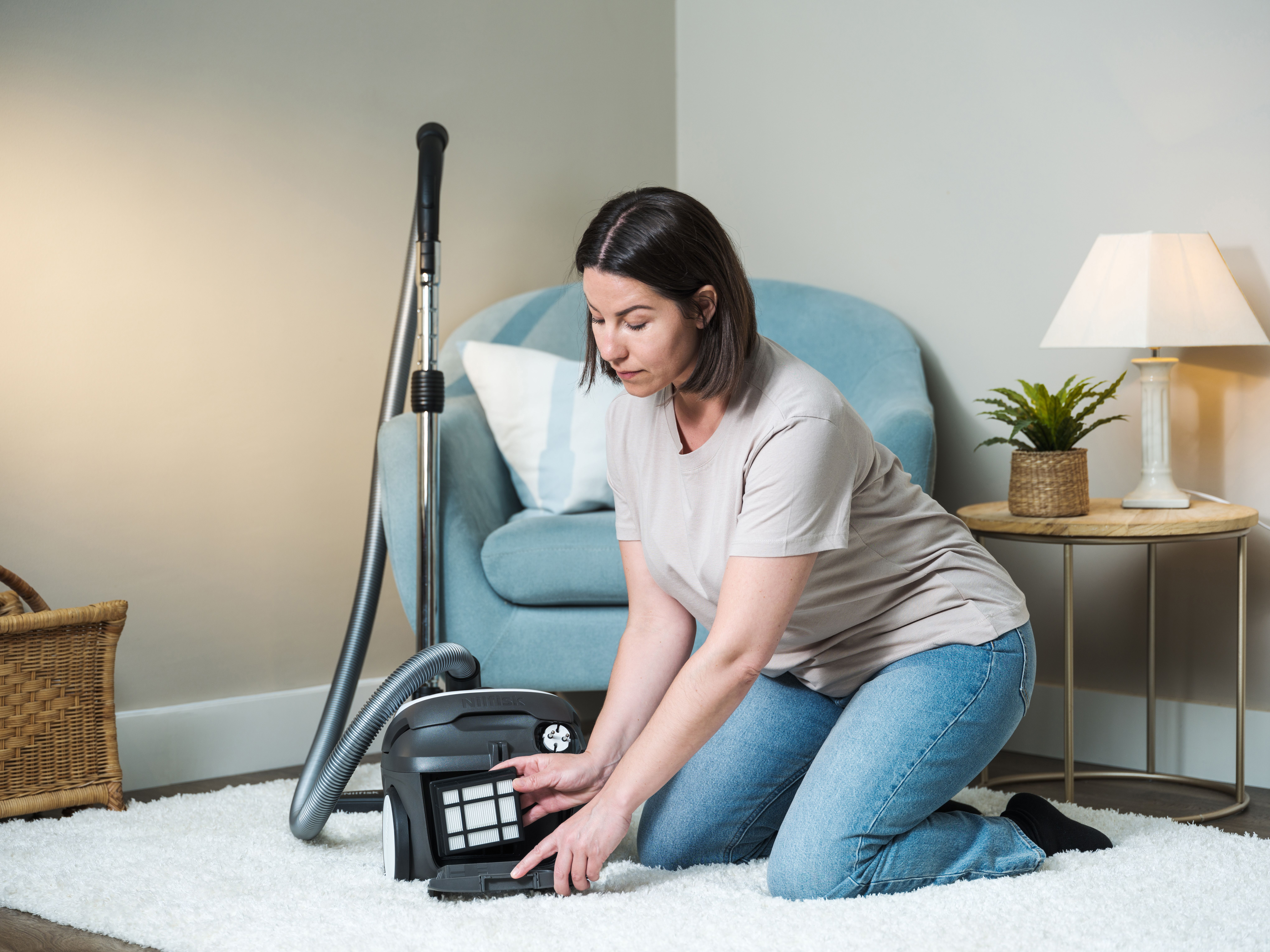Kobieta klęczy na białym dywanie, czyszcząc odkurzacz. W tle krzesło, lampa i kosz.