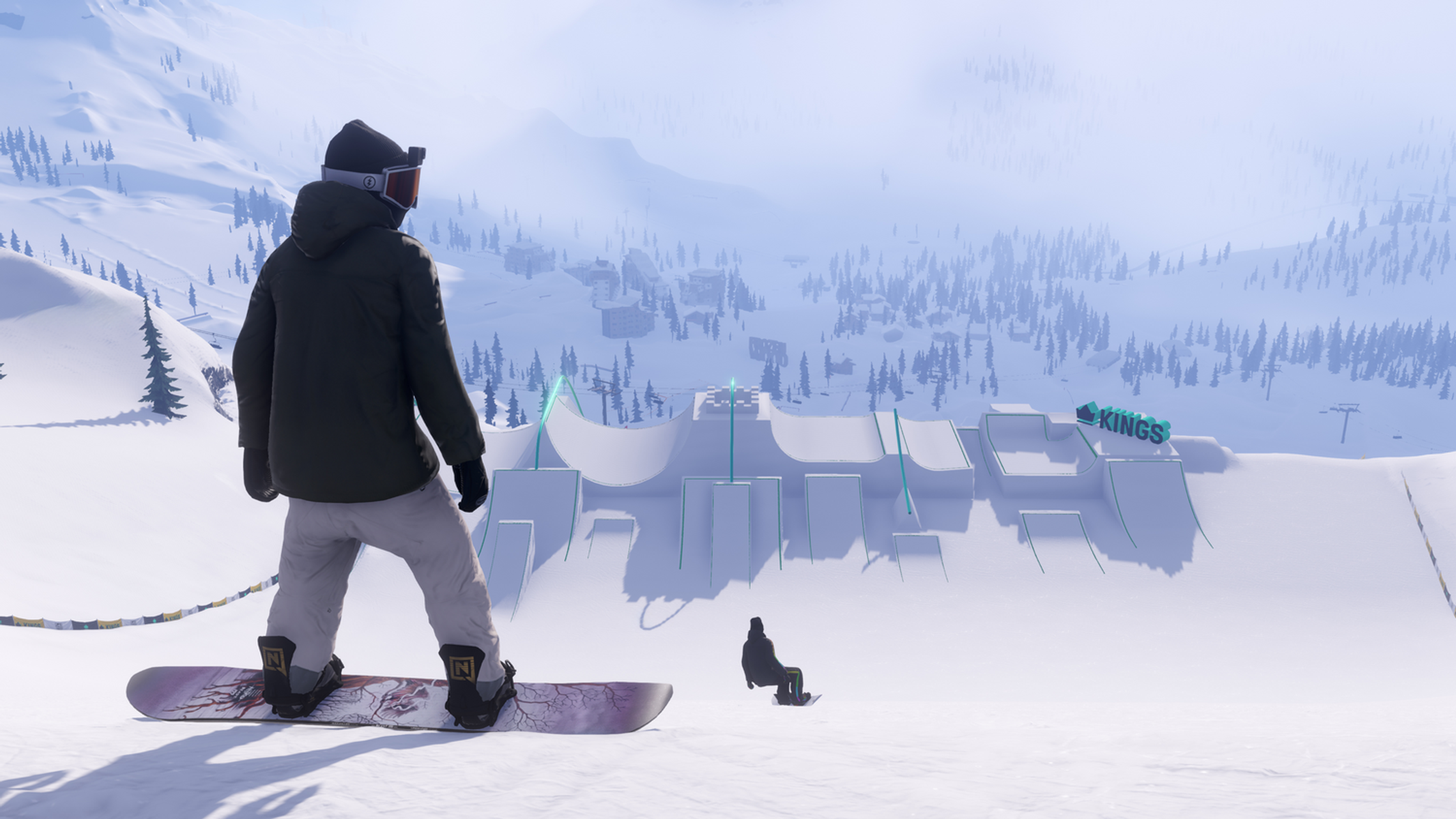 Uno snowboarder è in piedi su una pista innevata, guardando uno snowpark. Edifici e montagne sono visibili sullo sfondo.