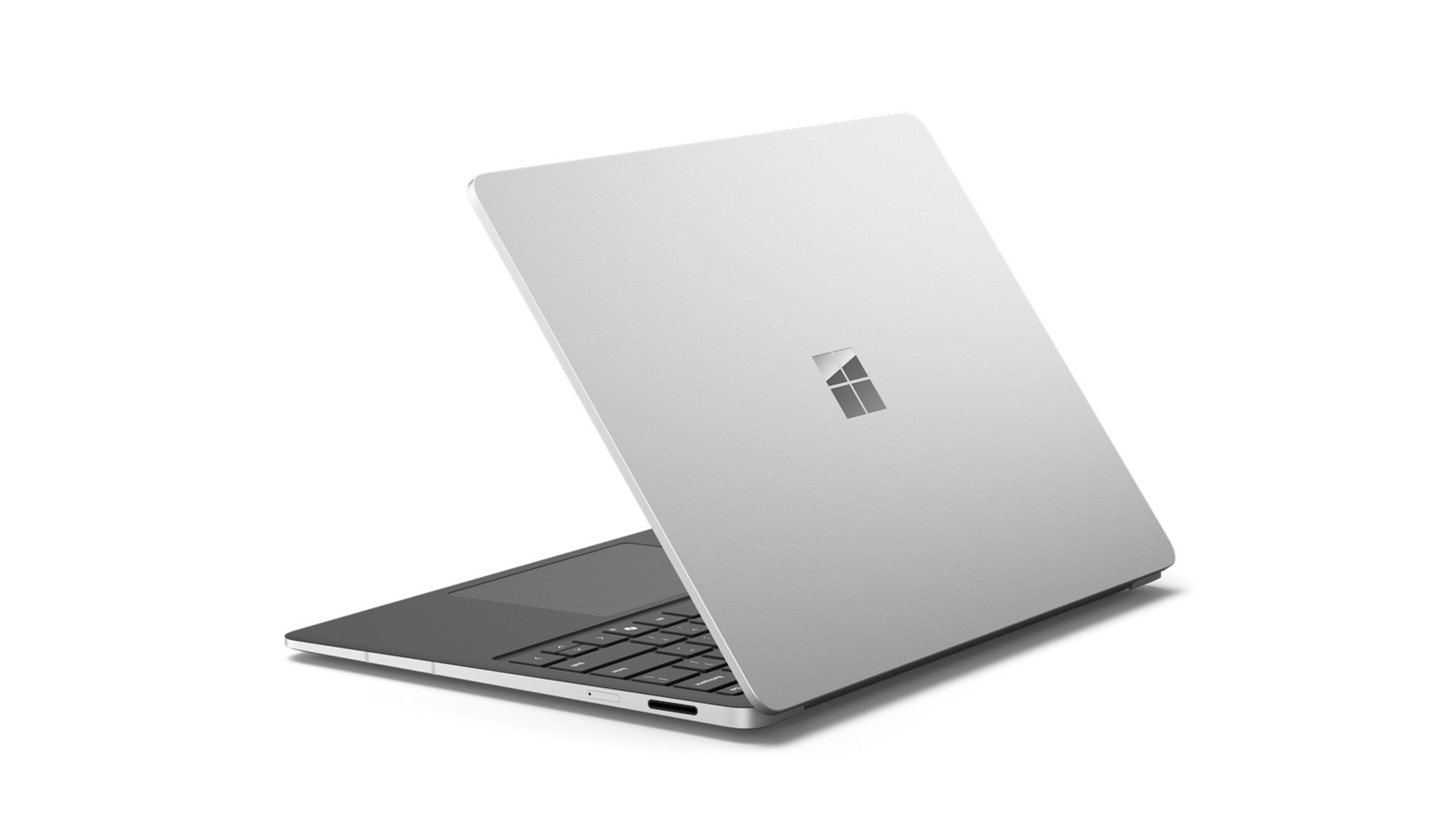 Un portátil plateado está abierto, mostrando el teclado y una pantalla con el logotipo de Microsoft. El fondo es negro.