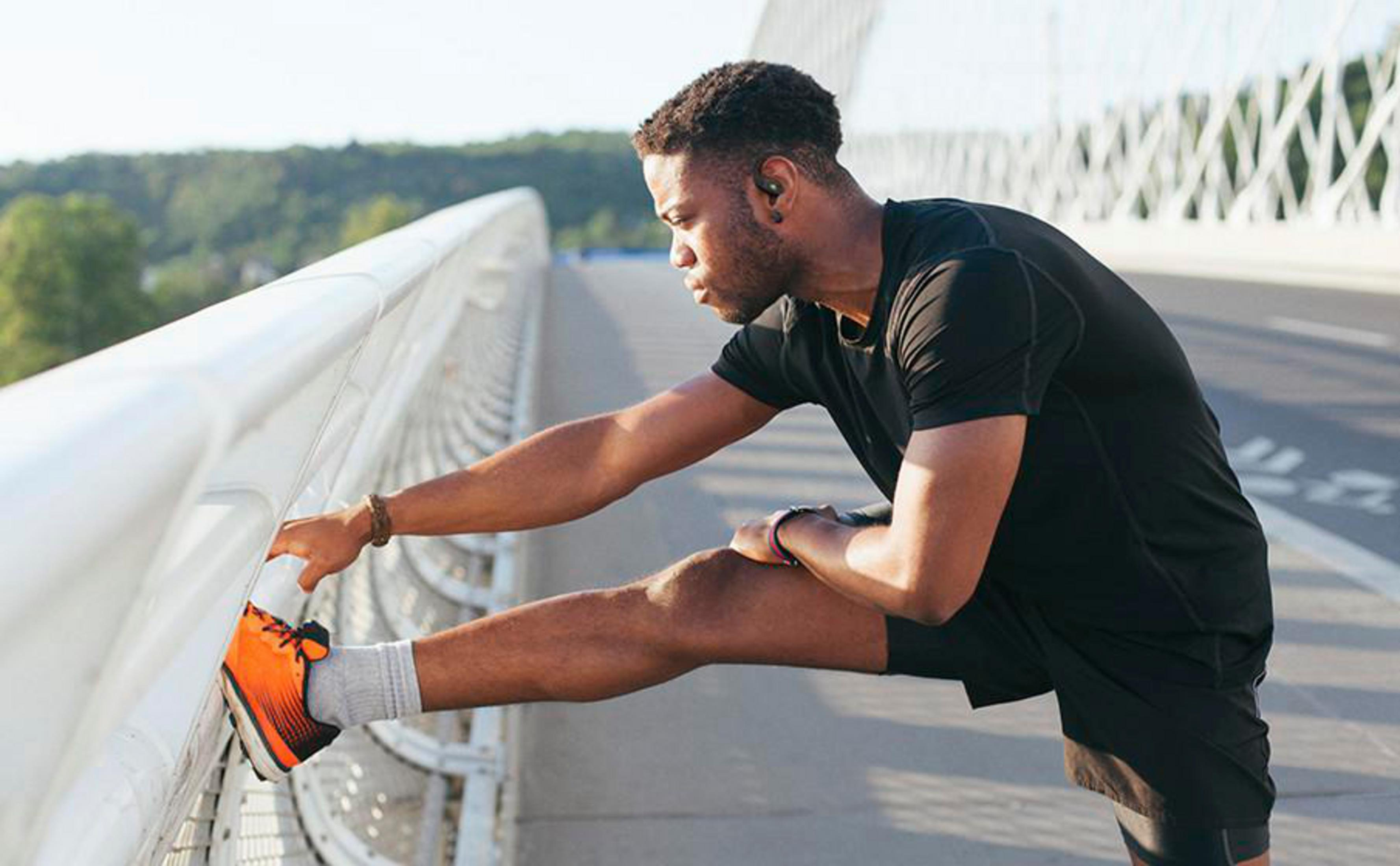 Uomo in nero si allunga su un ponte, indossa scarpe da ginnastica arancioni. Alberi verdi sullo sfondo.