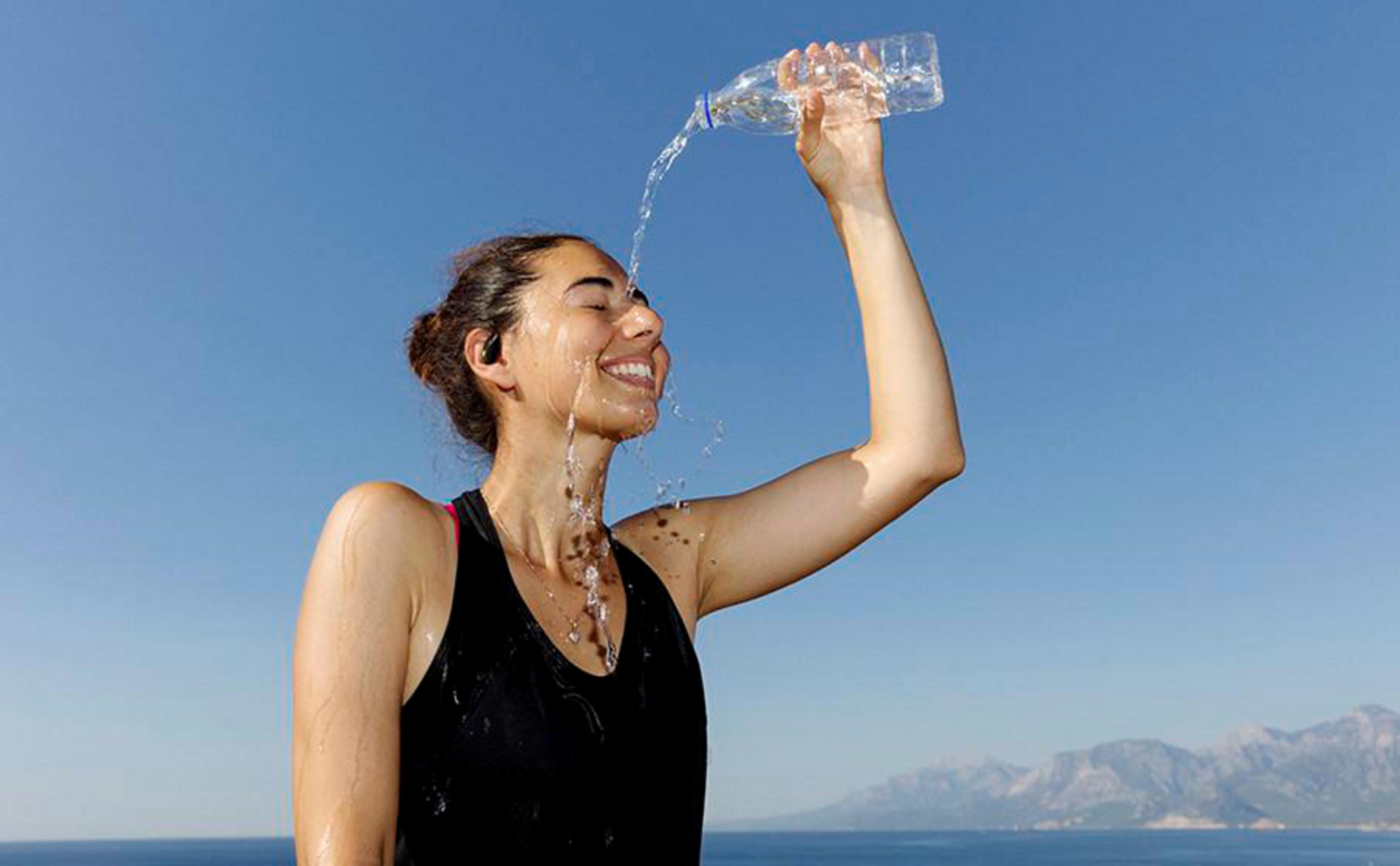 Una donna si versa l'acqua in faccia da una bottiglia. Sorride, con cielo blu, mare e montagne sullo sfondo.