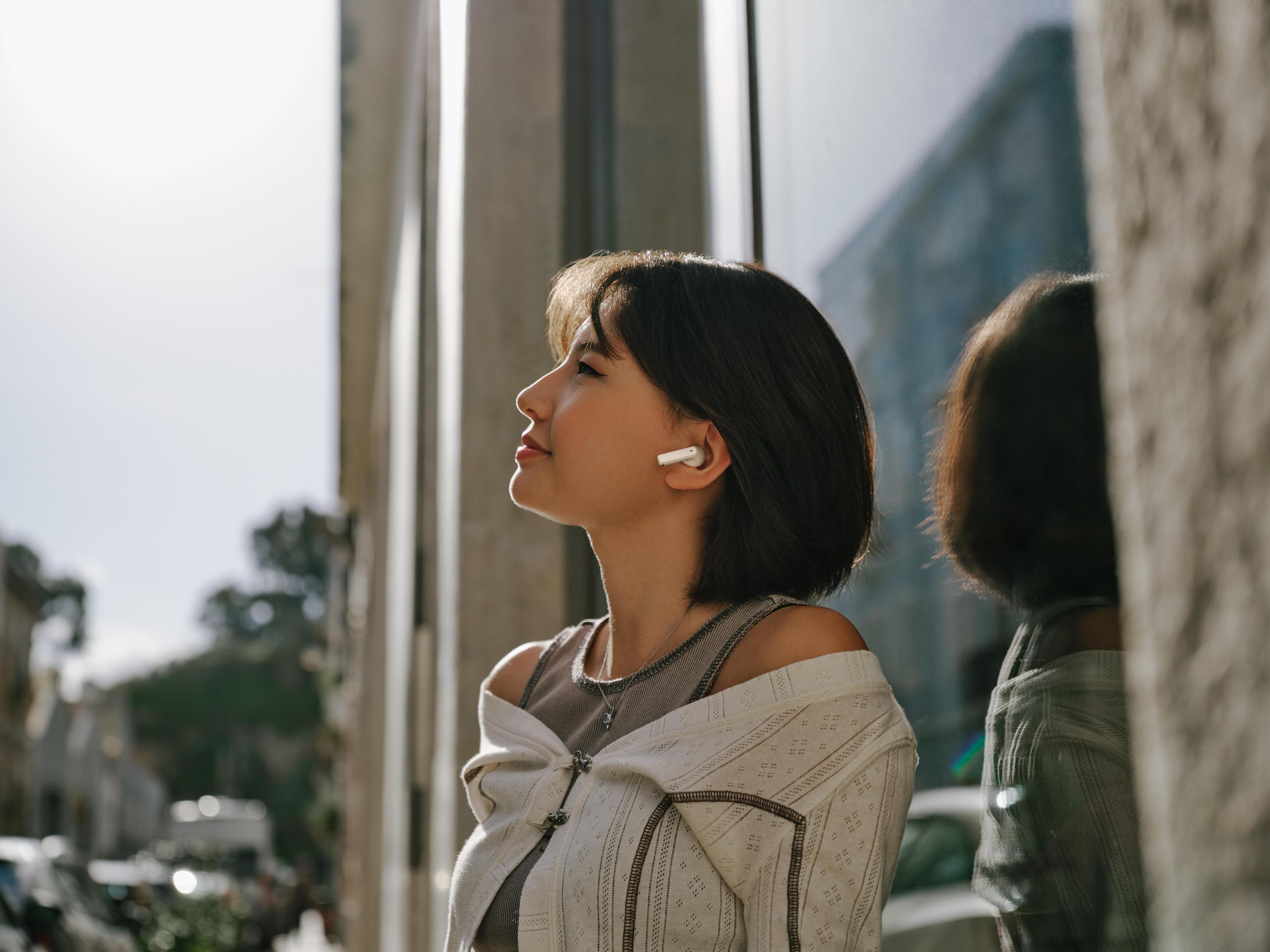 Mujer con cabello oscuro corto usando auriculares, mirando hacia arriba. Lleva un suéter claro. Se ve un reflejo en la ventana.
