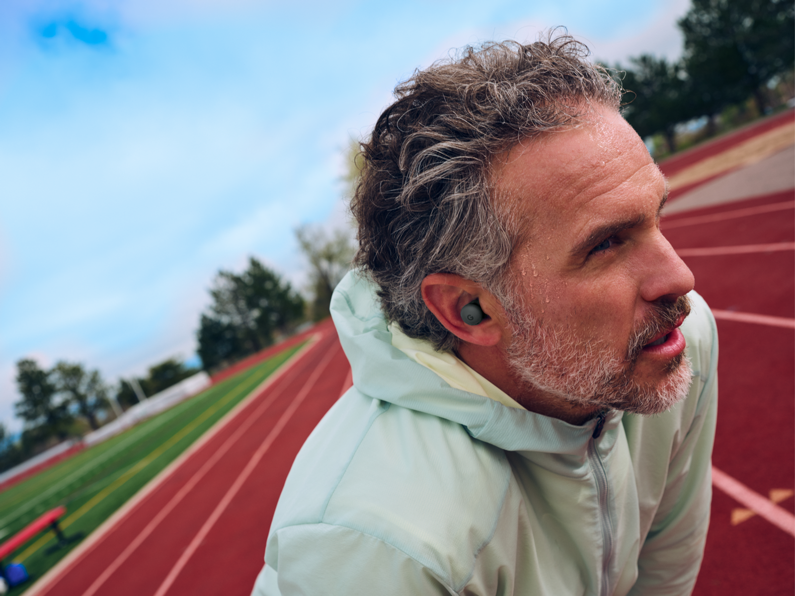 Man with gray hair and beard wearing earbuds, on a track. He is wearing a light green jacket, with a blue sky in the background.