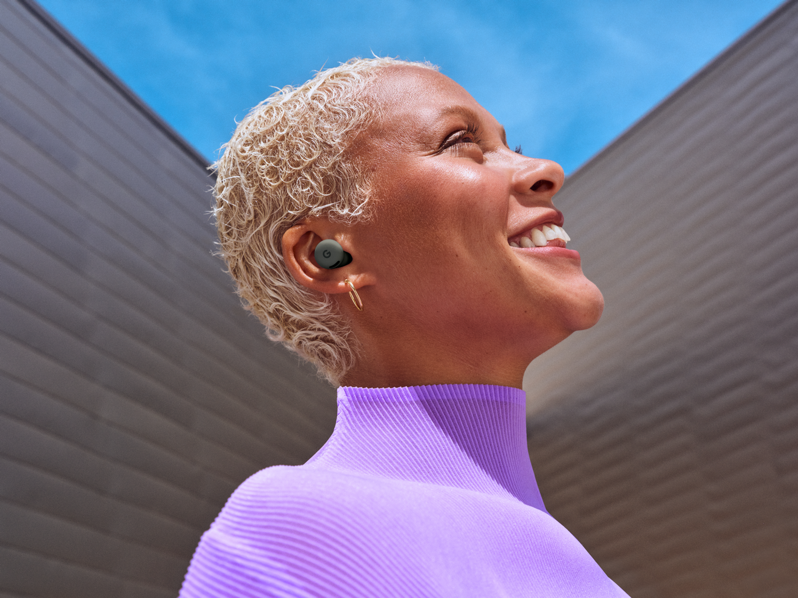A smiling person with short blonde hair wearing an earbud looks up. The background is blue sky and a gray building.
