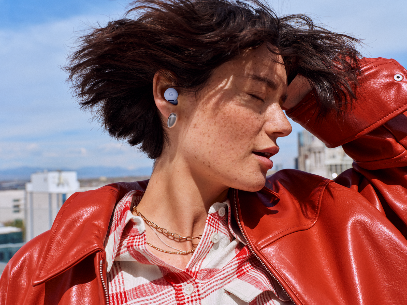 Eine Frau mit Sommersprossen, die eine rote Jacke und Ohrhörer trägt und nach oben blickt. Der Hintergrund hat einen blauen Himmel und ein Gebäude.