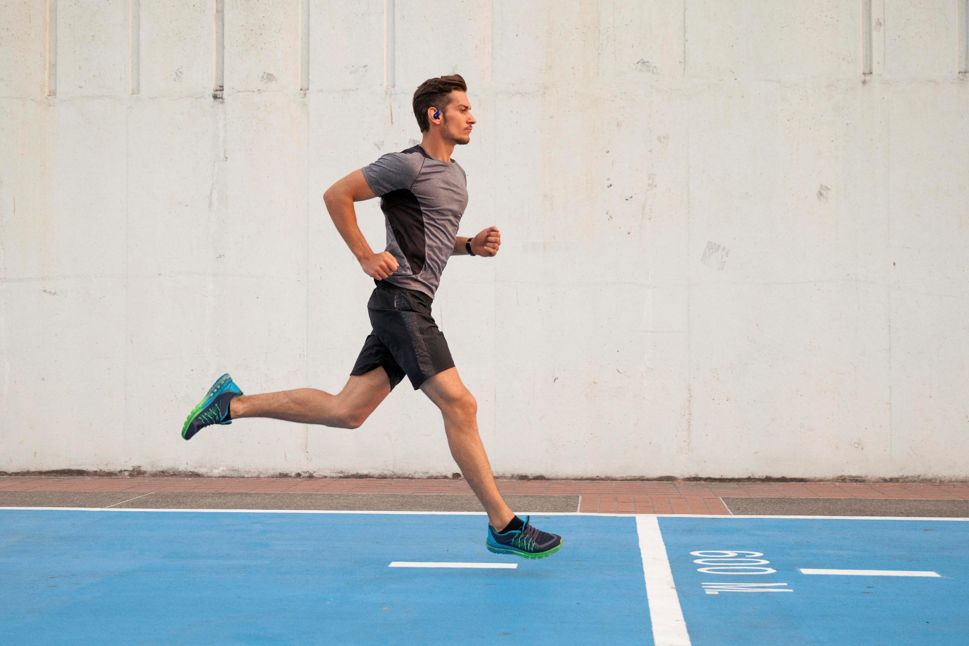 Uomo che corre all'aperto. Indossa maglia grigia, pantaloncini neri e scarpe blu/verdi. Superficie blu e muro bianco sullo sfondo.