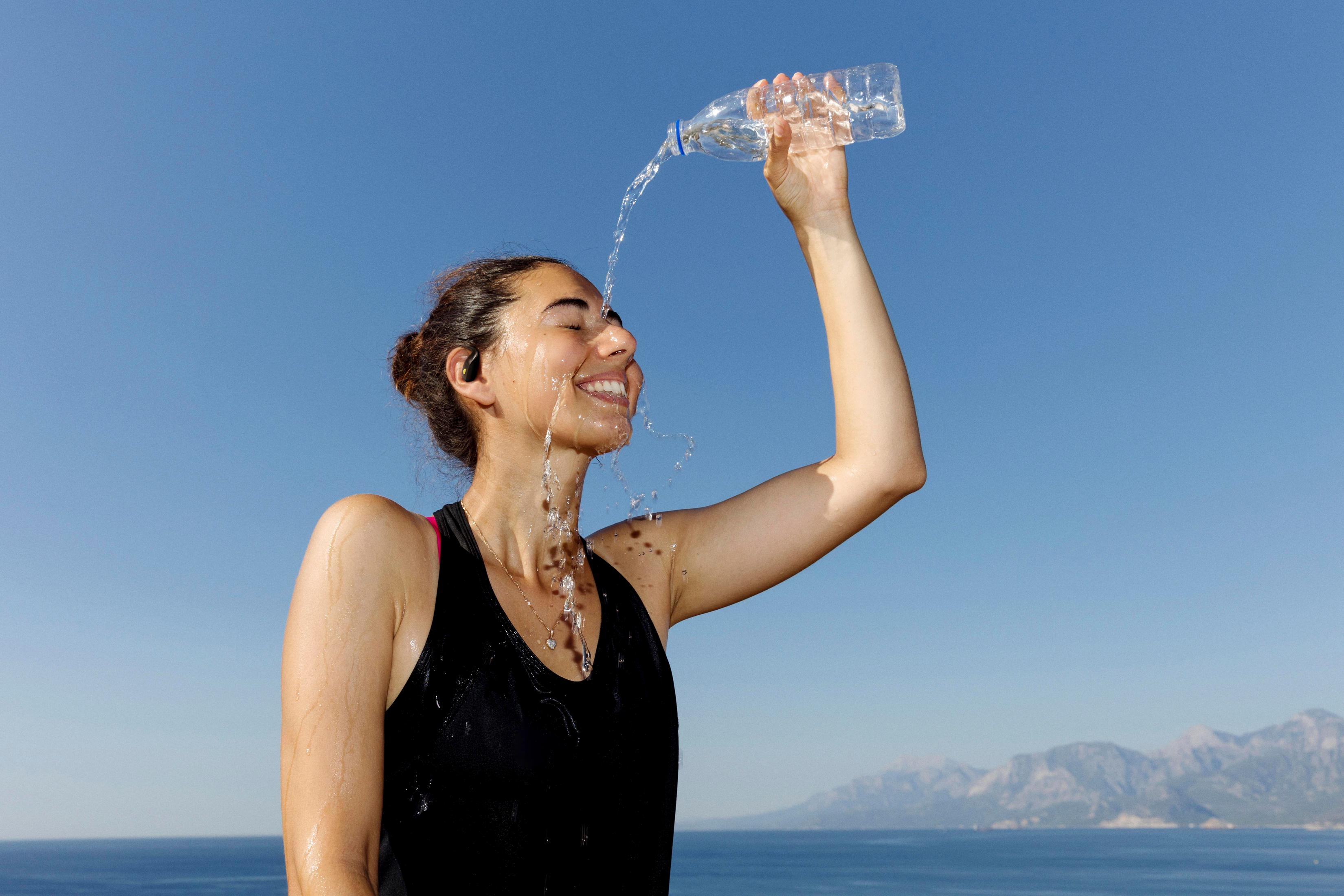 Una donna si versa acqua sul viso. Indossa un top nero e sorride. Cielo blu e mare sullo sfondo.