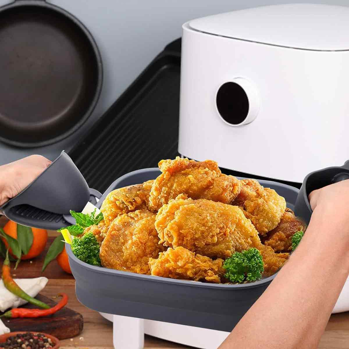 Eine Person hält ein Tablett mit gebratenem Hähnchen, im Hintergrund ein weißes Gerät. Weitere Küchenutensilien auf einer Holzoberfläche sichtbar.