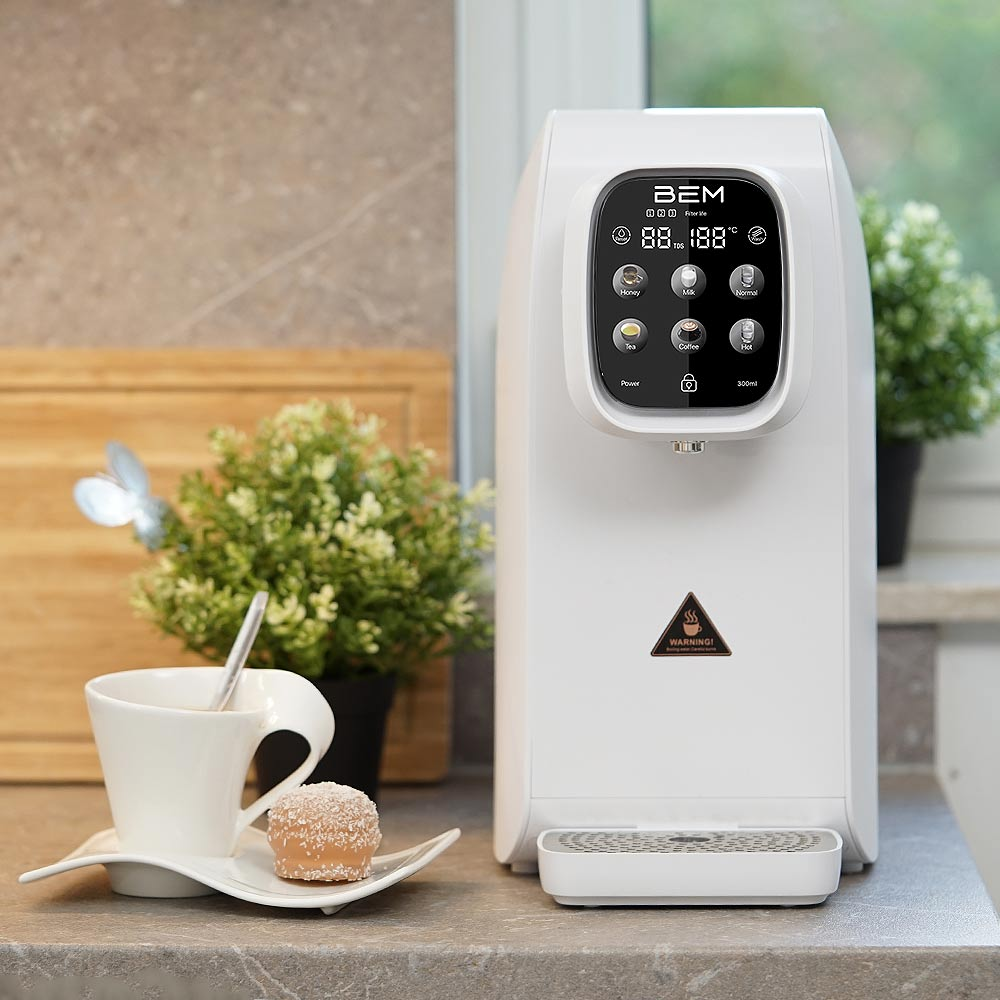 Ein weißer Wasserspender mit Digitalanzeige, eine Tasse Tee und ein kleiner Kuchen. Grüne Pflanzen im Hintergrund.