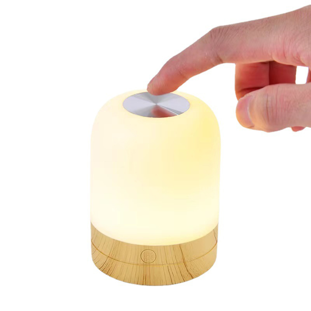 Eine Hand berührt eine leuchtende Lampe. Die Lampe ist zylindrisch mit Holzsockel. Die Oberseite ist silber. Der Hintergrund ist weiß.