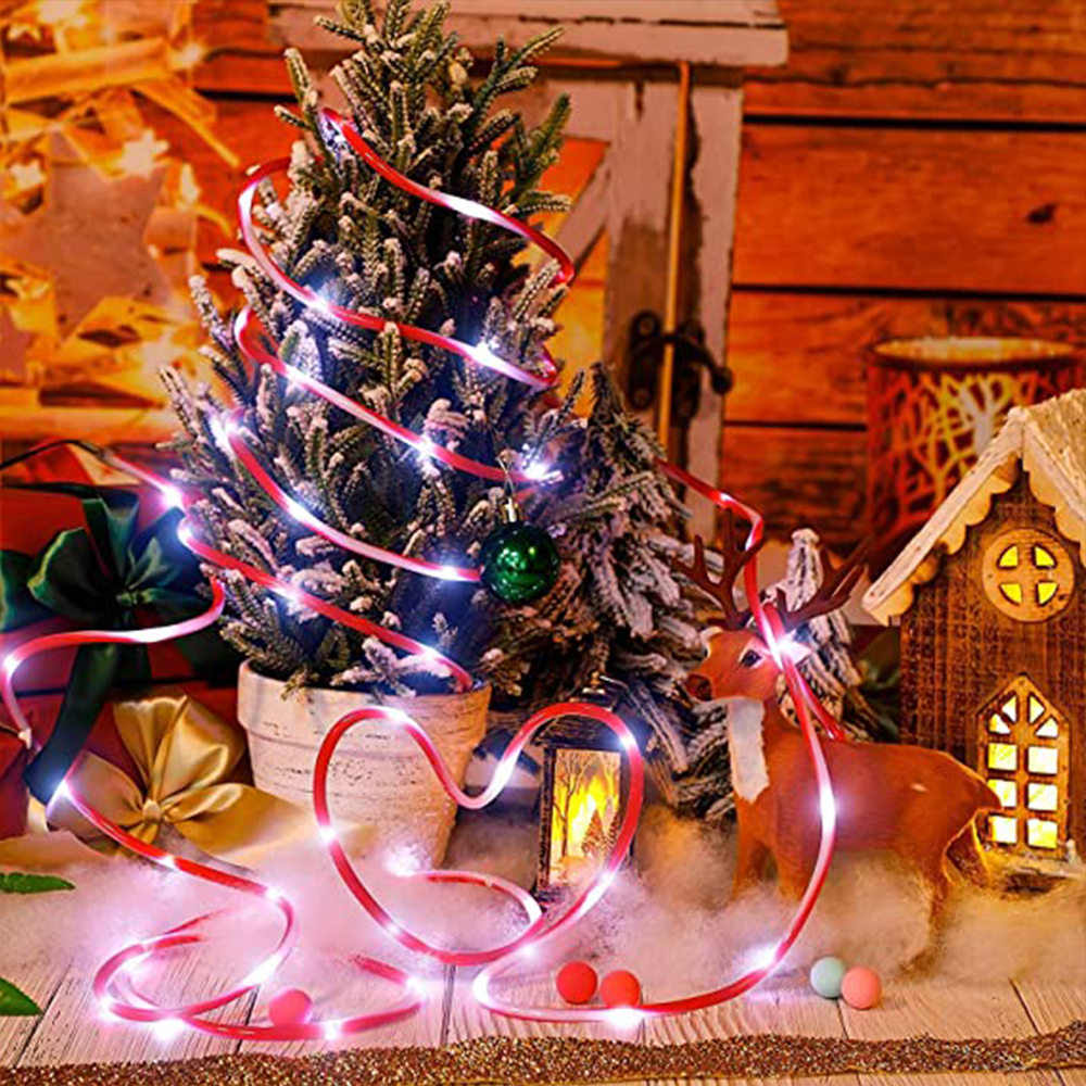 Weihnachtsszene mit geschmücktem Baum, Lichtern, Hirsch und kleinem Haus. Rotes Band mit Lichtern bildet Herzform.
