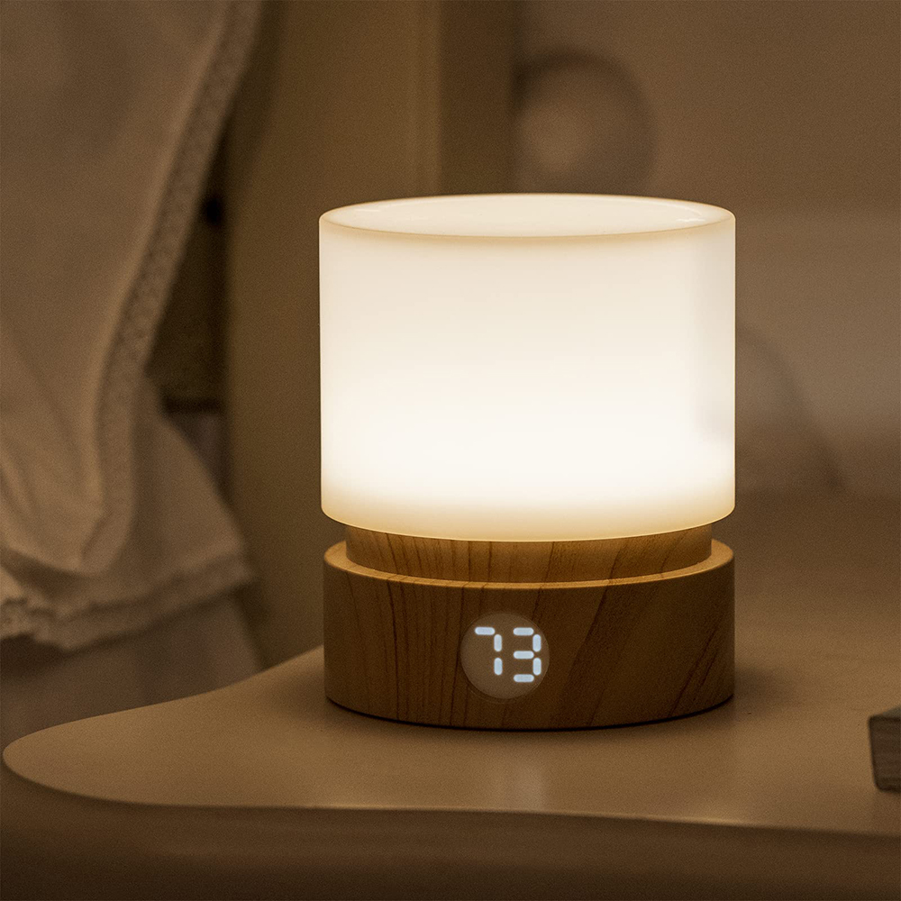 Eine beleuchtete Nachttischlampe mit Holzsockel, die "73" anzeigt. Die Lampe ist zylindrisch und strahlt sanft, auf einem Nachttisch.