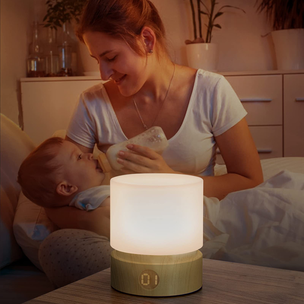 Eine Mutter füttert ein Baby mit einer Flasche. Auf dem Nachttisch steht eine Lampe mit Holzsockel und leuchtender Oberseite. Die Szene ist drinnen.