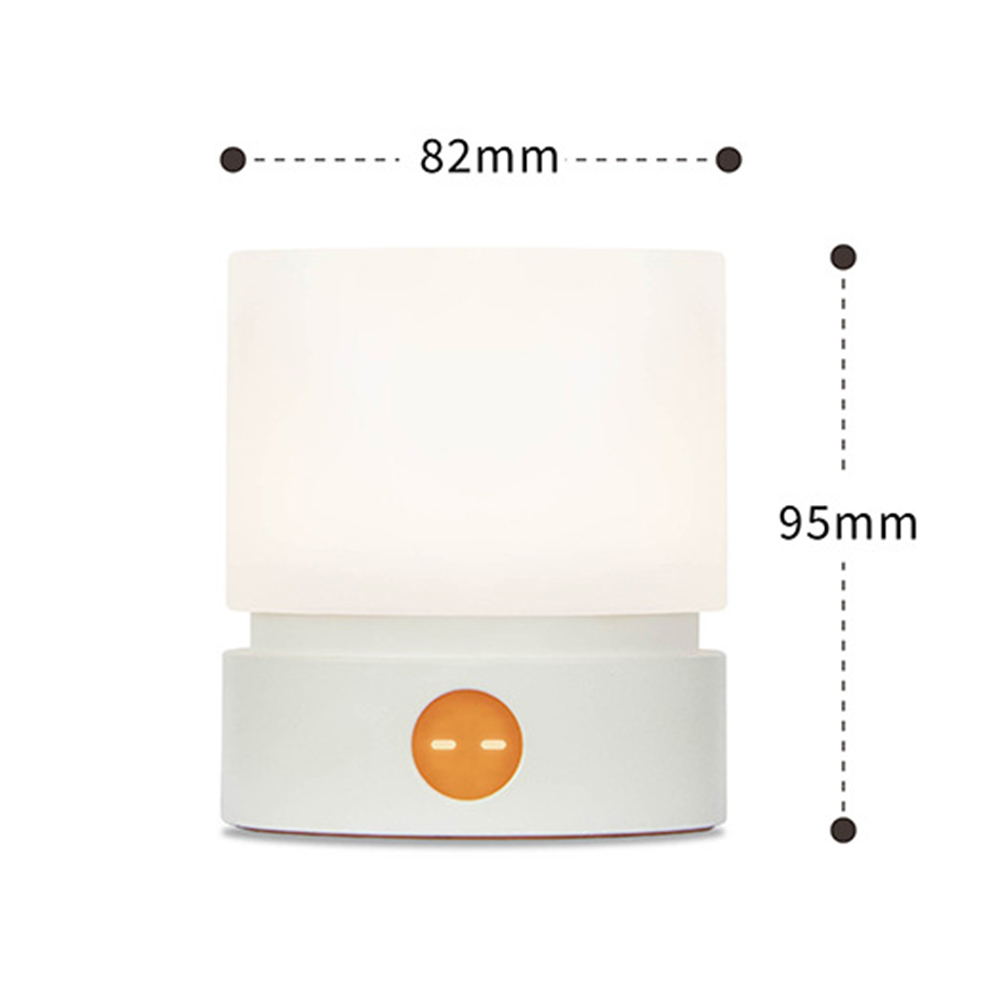 Eine kleine, weiße Nachttischlampe mit orangefarbenem Knopf. Sie ist 82 mm breit und 95 mm hoch. Die Lampe leuchtet und gibt ein weiches Licht ab.