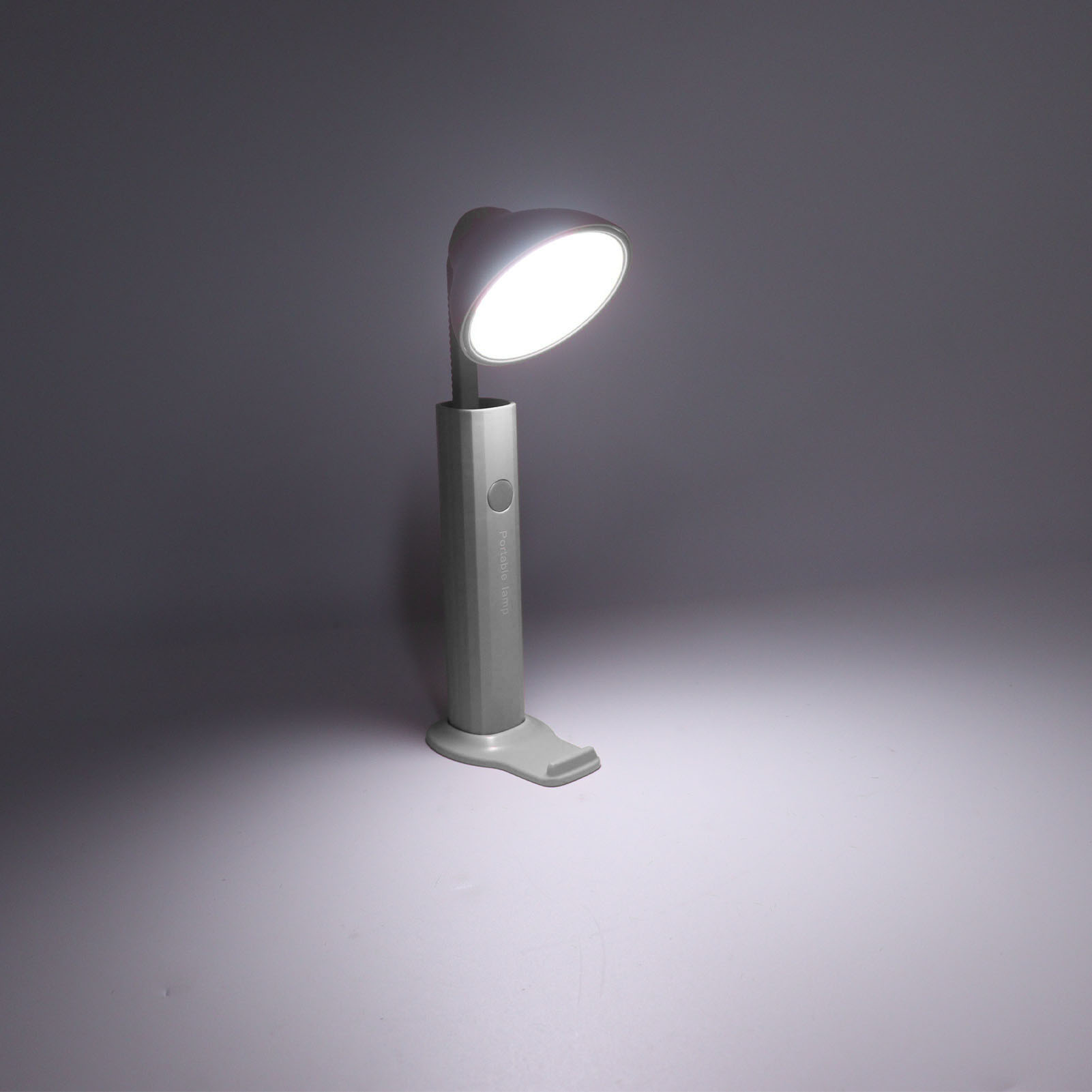Eine graue tragbare Lampe steht auf einer weißen Oberfläche. Die Lampe leuchtet und wirft ein helles Licht. Der Hintergrund ist dunkelgrau.
