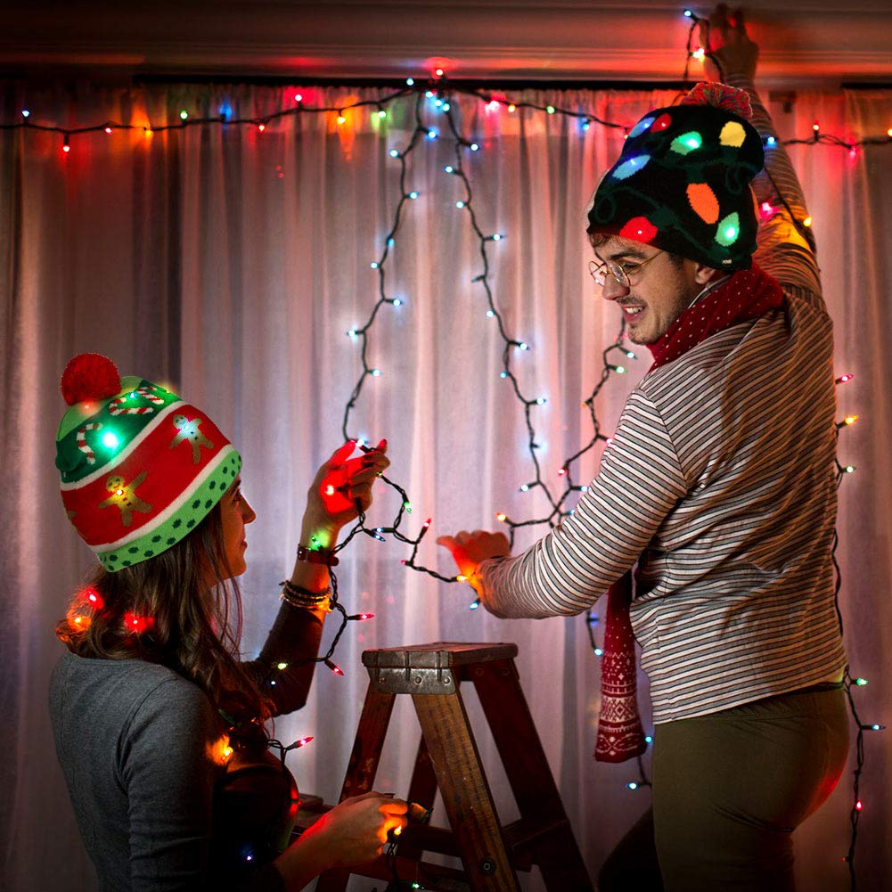 Ein Paar dekoriert mit Weihnachtsbeleuchtung. Sie tragen festliche Hüte und stehen in der Nähe einer Leiter. Der Hintergrund ist ein Vorhang mit mehr Lichtern.