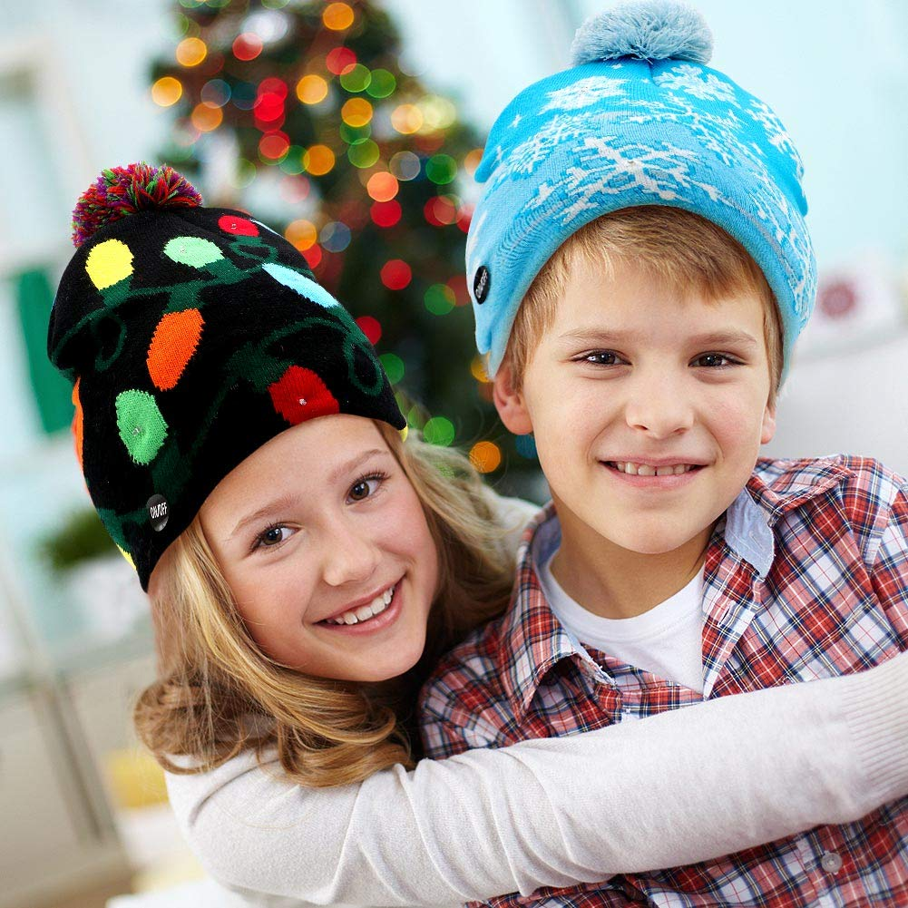Zwei lächelnde Kinder tragen Wintermützen. Eine schwarze mit Lichtern, die andere blau mit Schneeflocken. Im Hintergrund ein Weihnachtsbaum.