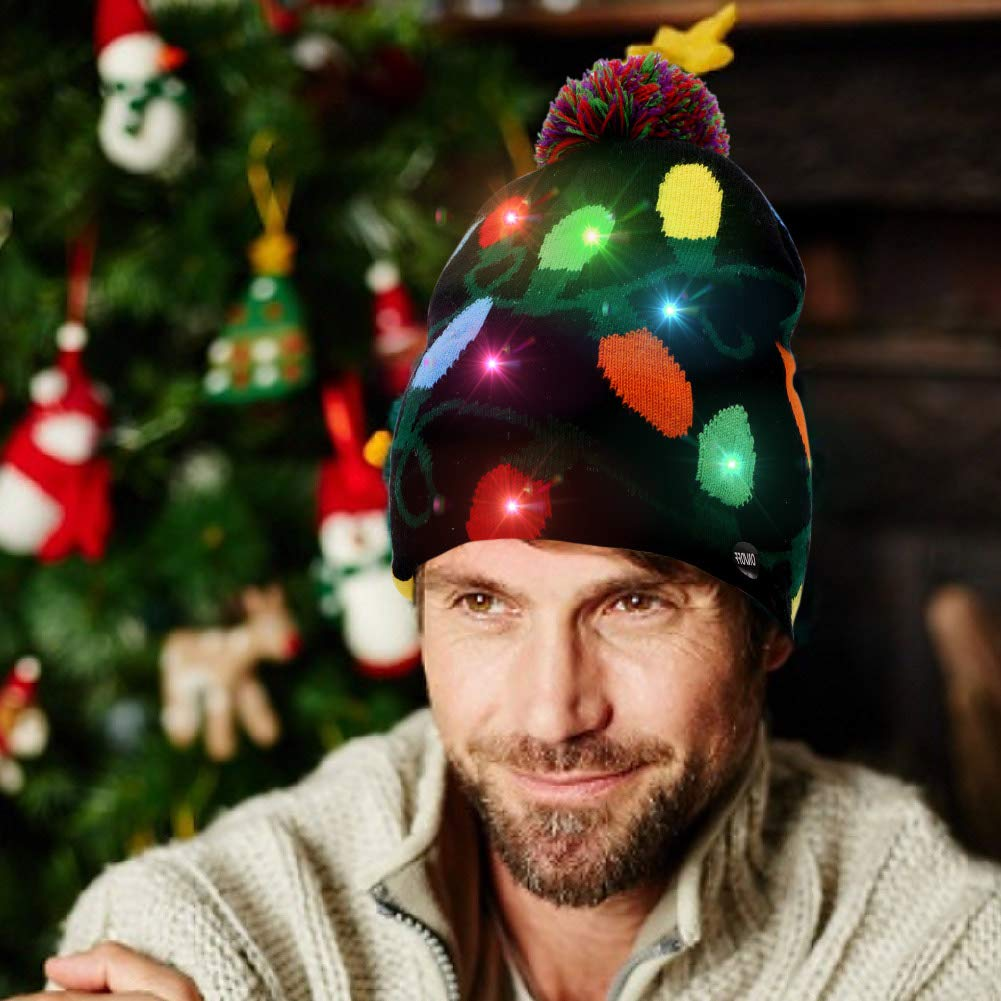 Ein Mann trägt eine schwarze Mütze mit bunten Lichtern vor einem Weihnachtsbaum. Er lächelt.