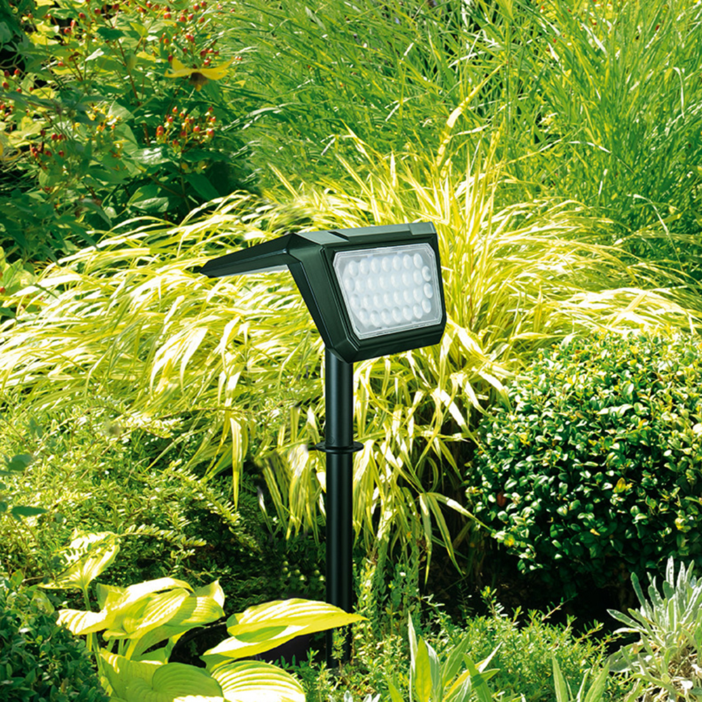 Eine schwarze LED-Gartenleuchte steht inmitten grüner und gelber Pflanzen. Das Licht ist an und beleuchtet das umliegende Laub.