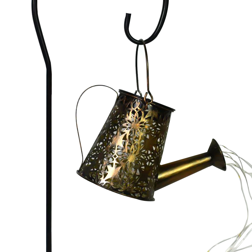 Eine bronzefarbene Gießkanne mit dekorativen Ausschnitten hängt. Weiße Lichter strahlen aus dem Ausguss. Ein schwarzer Metallstab stützt die Kanne.
