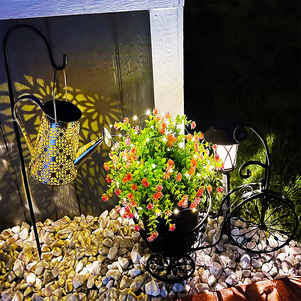 Eine dekorative Gartenszene bei Nacht. Mit Blumenarrangement in einem Fahrradbehälter, einer hängenden Gießkanne und einer Lampe.