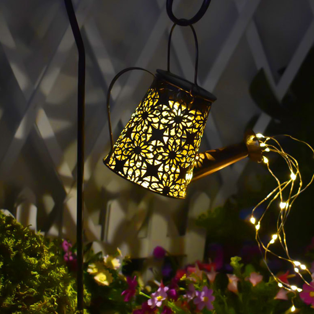 Eine dekorative Gießkanne-Laterne hängt und ist mit warmen Lichtern beleuchtet. Blumen und Laub befinden sich im Vordergrund. Dahinter ist ein weißer Zaun.