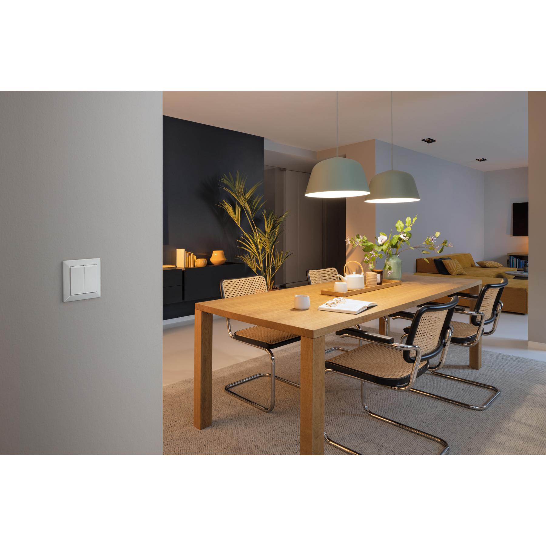 Jadalnia ze stołem, krzesłami i lampami wiszącymi. Włącznik światła na ścianie. Sofa w tle.