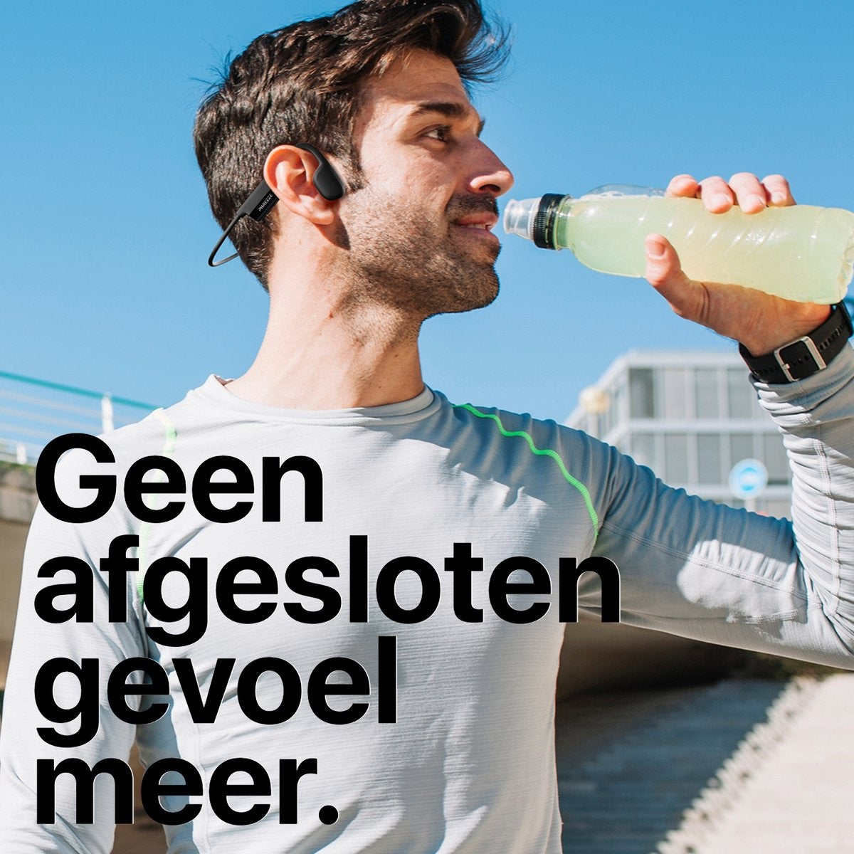 Man met koptelefoon drinkt uit een fles. Hij draagt ​​een grijs shirt en een zwart horloge. Helderblauwe lucht op de achtergrond.