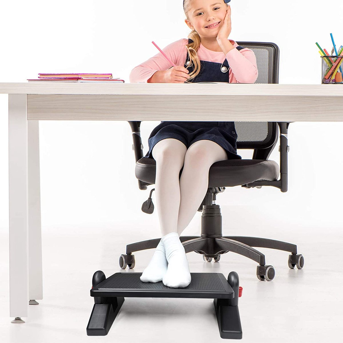 A young girl sits at a desk, smiling. She has a pencil in hand. White socks and a footrest under the desk.