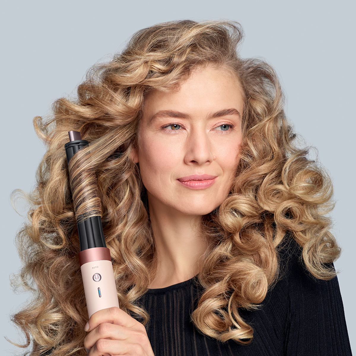 Kobieta z blond kręconymi włosami kręci pasmo loków stylizatorem. Ma na sobie czarny top i jasną skórę. Tło jest szare.