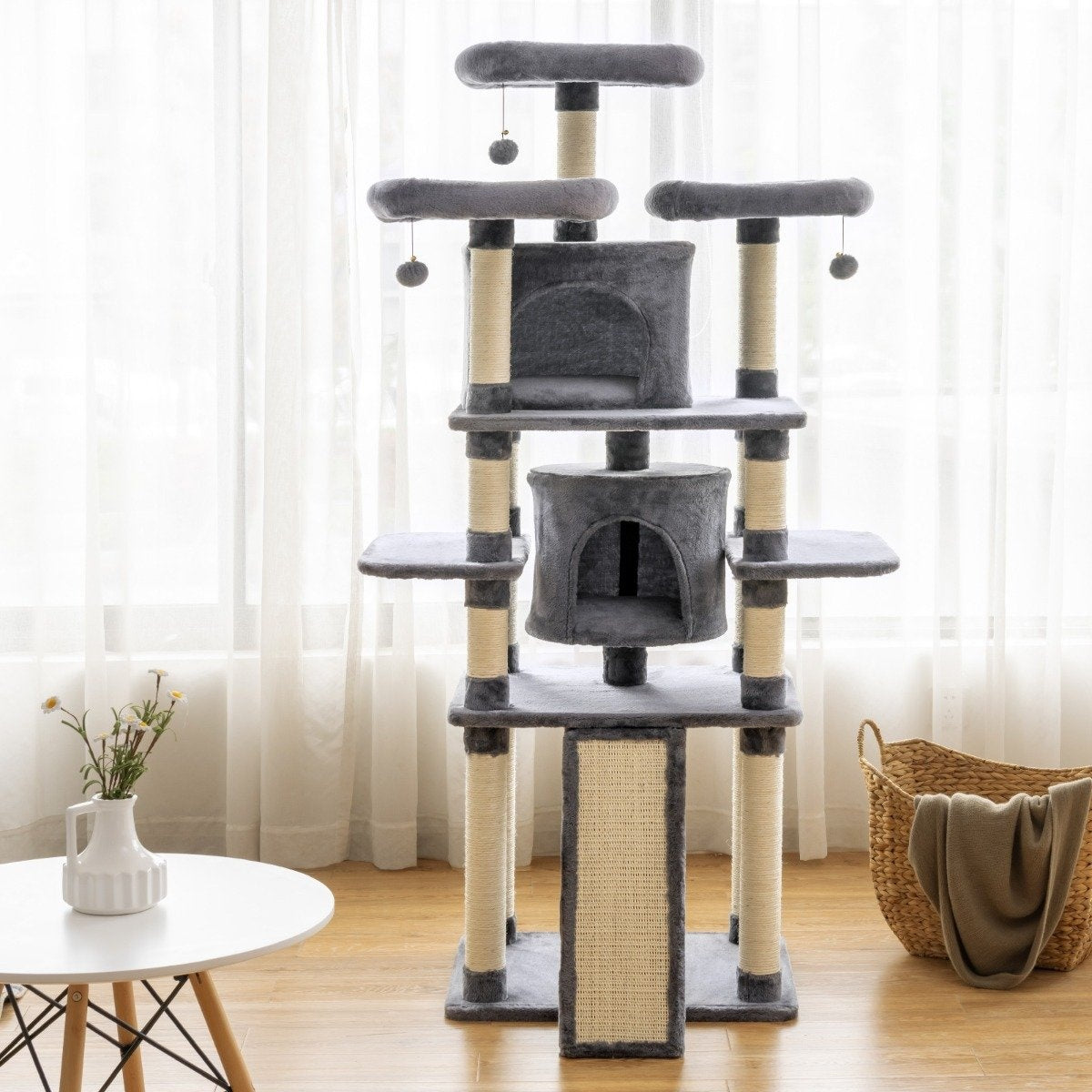 Un árbol para gatos gris con múltiples plataformas, escondites y postes para rascar. Una mesa blanca con flores y una cesta delante.