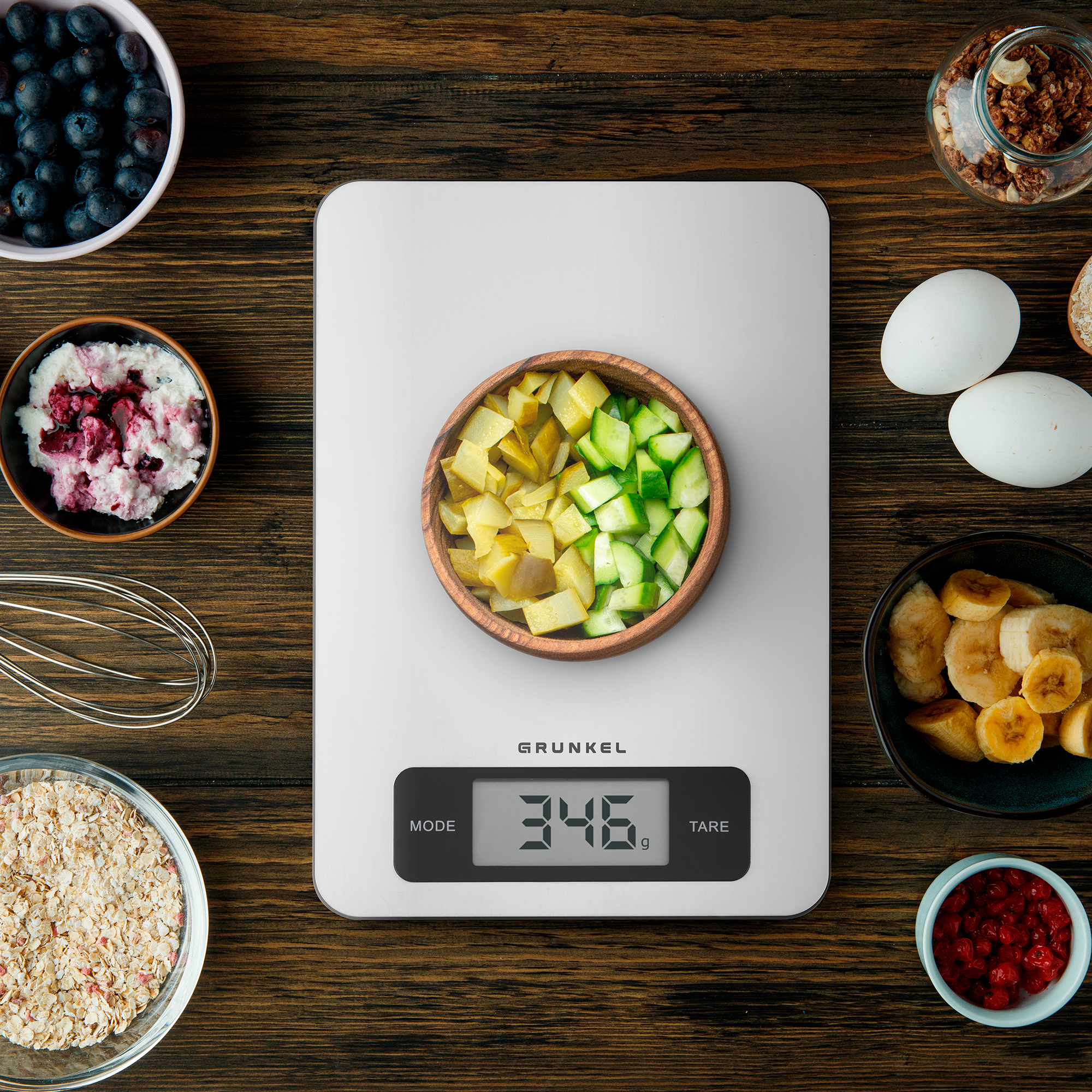 Mesa de madera con ingredientes, una báscula digital que muestra 346g y cuencos con bayas y fruta. Vista superior.