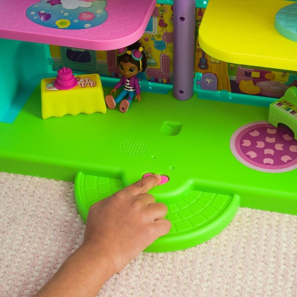 Die Hand eines Kindes drückt einen rosa Knopf auf einem grünen Spielzeughaus. Eine Puppe sitzt auf der grünen Basis. Bunte Ebenen und Zubehör sind vorhanden.