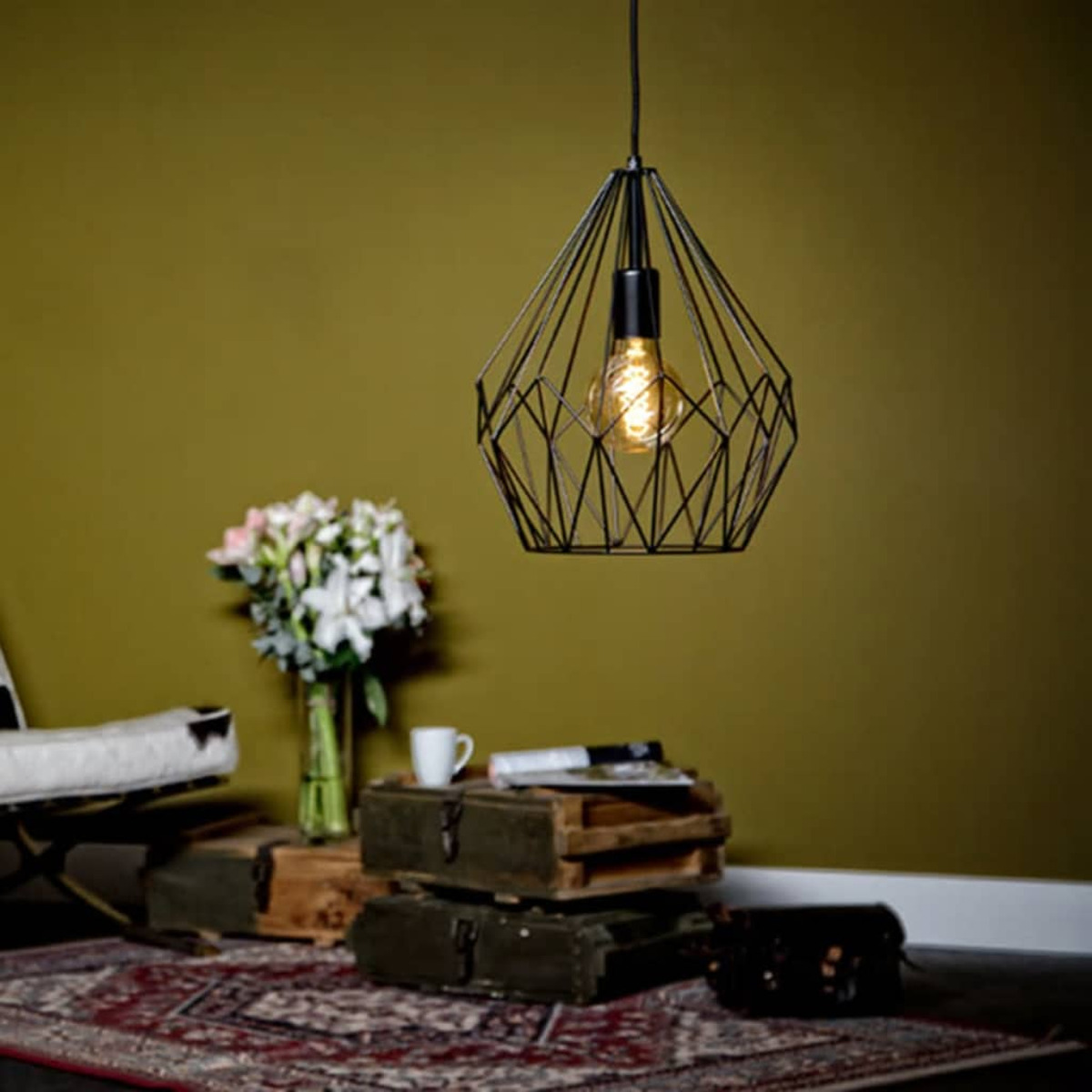 Eine geometrische schwarze Pendelleuchte hängt über Holzkisten, Blumen und einem Teppich. In der Leuchte befindet sich eine Glühbirne. Grüne Wand.