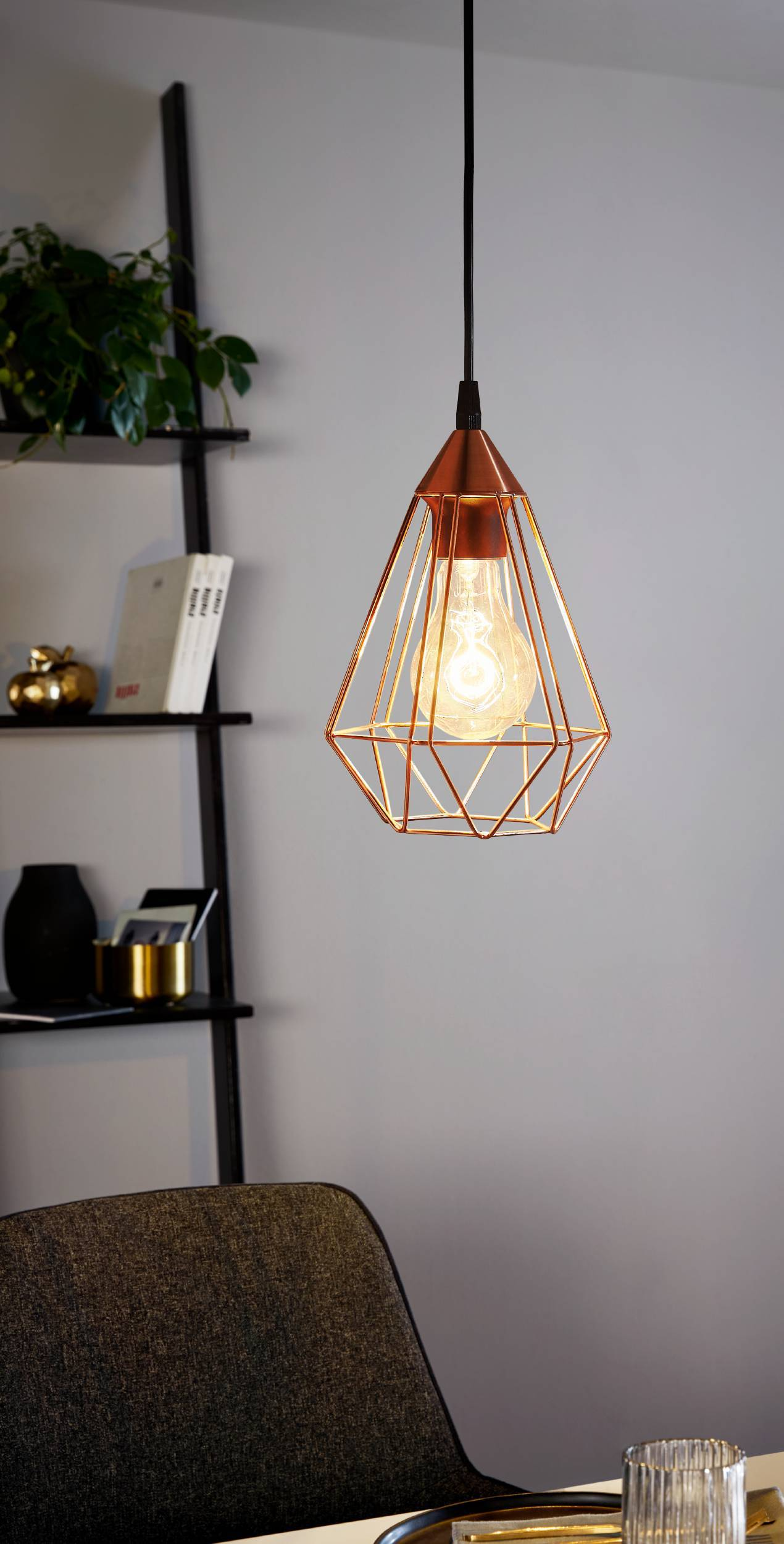 Eine kupferfarbene geometrische Pendelleuchte beleuchtet einen Raum. Im Hintergrund ein Regal mit Büchern und Pflanzen, im Vordergrund ein Stuhl.